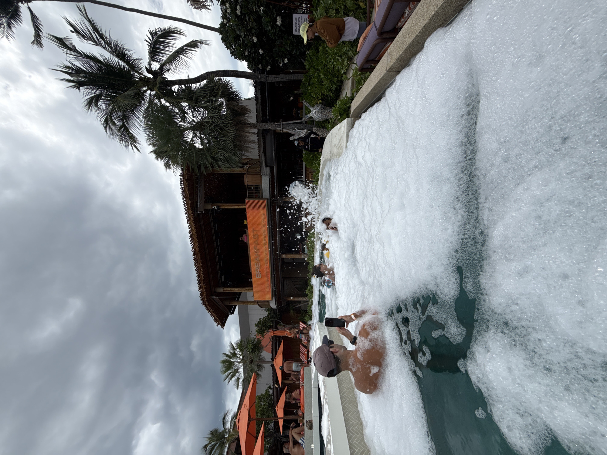 In Ko Samui District, Thailand, a foam party is taking place in a swimming pool. The pool is filled with a thick layer of white foam, obscuring much of the water. In the foreground, a man wearing a baseball cap and sunglasses is partially submerged in the foam, holding a smartphone to take a photo. Behind him, other people are visible in the foam, some interacting with each other. In the background, there is a building with a thatched roof and balconies, likely a bar or restaurant, with an orange sign that reads "BREAKFAST" and other text below it. Palm trees are scattered throughout the scene, and the sky is overcast with dramatic clouds, suggesting a humid, tropical climate. Orange umbrellas are visible in the distance, indicating a resort or beach club setting. A woman in a yellow hat and brown jacket stands on the edge of the pool area, observing the scene. The overall atmosphere appears to be lively and recreational.