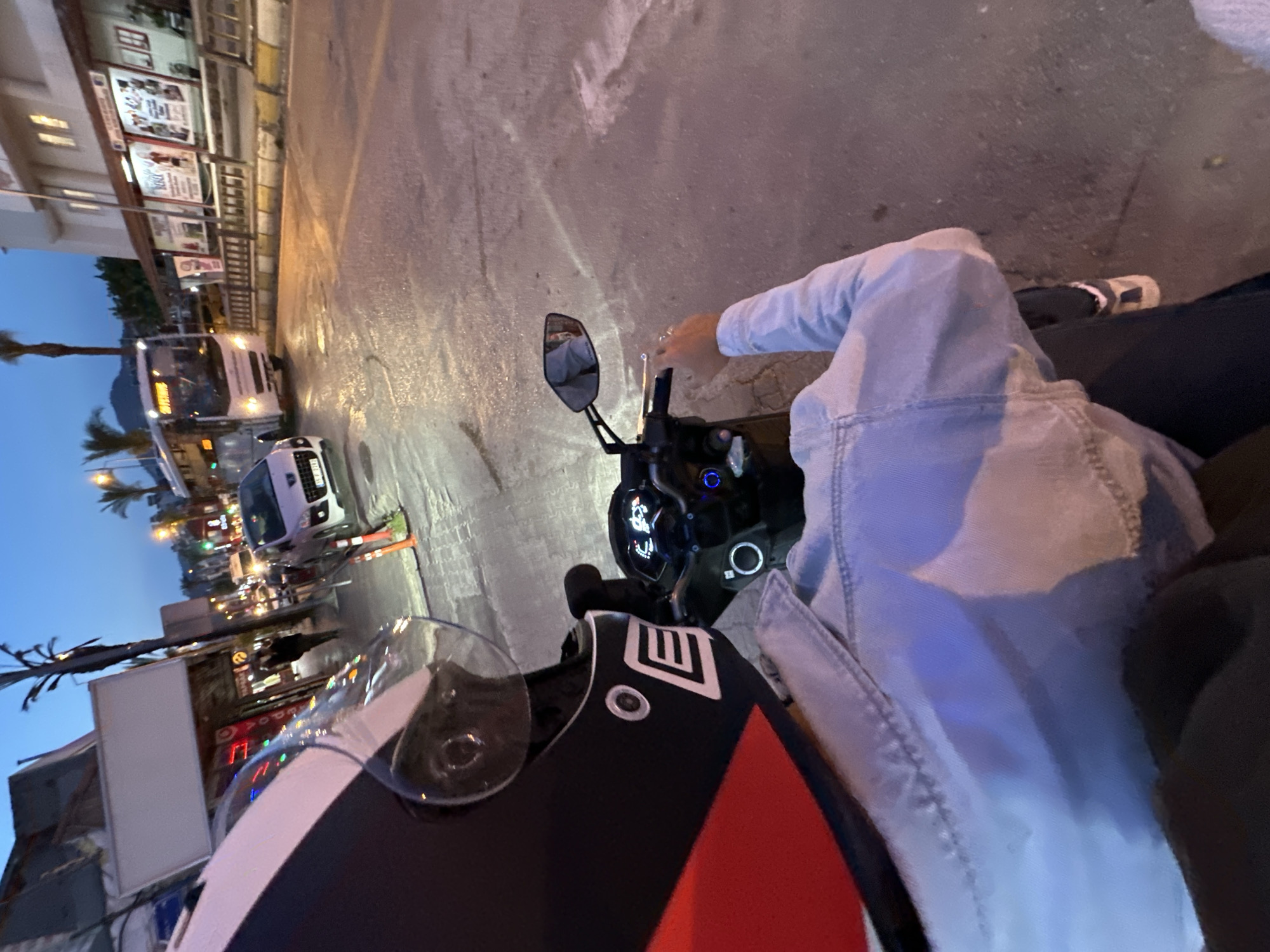 The image shows a first-person view from a person riding a motorcycle on a street in Fethiye, Turkey. The rider is wearing a helmet with a visor, a light blue denim jacket, and dark pants. The motorcycle has a black control panel and a side mirror. The street is paved and illuminated by streetlights and headlights from other vehicles. Buildings line the left side of the street, with signs and storefronts visible. A bus and several cars are driving on the road. Palm trees and other vegetation are also visible. The sky is a gradient of blue, indicating either dusk or dawn.