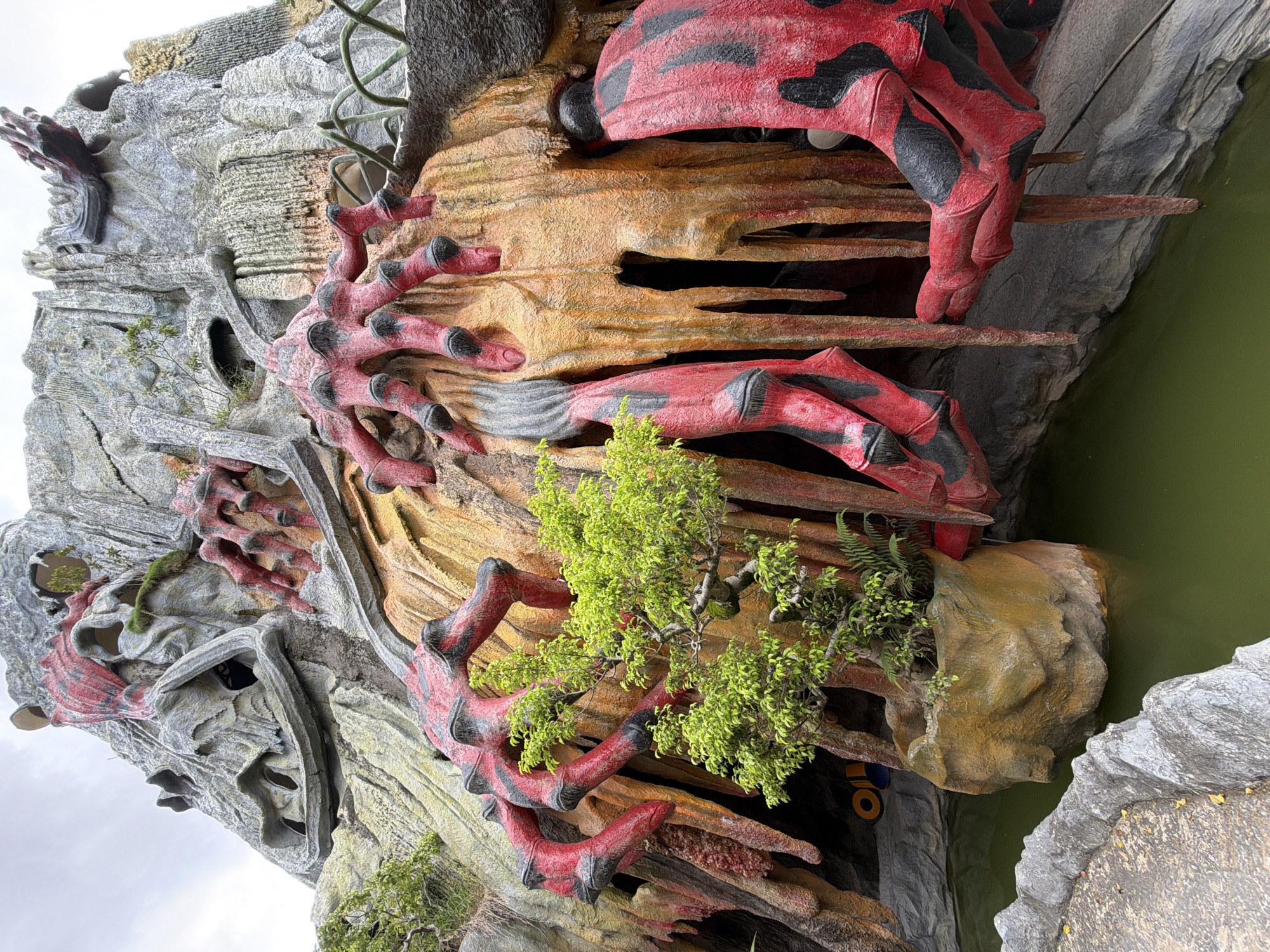 The image depicts a close-up view of a surreal, artificial rock formation resembling a monstrous creature. This structure, located at the Crazy House (also known as Hằng Nga Guesthouse) in Da Lat, Vietnam, is adorned with vibrant red and black appendages that appear to be grasping or emerging from the rocky surface. The rock itself is textured and colored in shades of grey, orange, and yellow, mimicking stalactites and organic forms. A small, lush green tree with delicate branches is planted in a rocky basin, contrasting with the alien aesthetic of the surrounding sculpture. Below the rock formation, a pool of murky green water reflects the unusual scenery. The sky above is overcast, suggesting a cloudy day. The perspective is from ground level, looking up at the imposing structure.