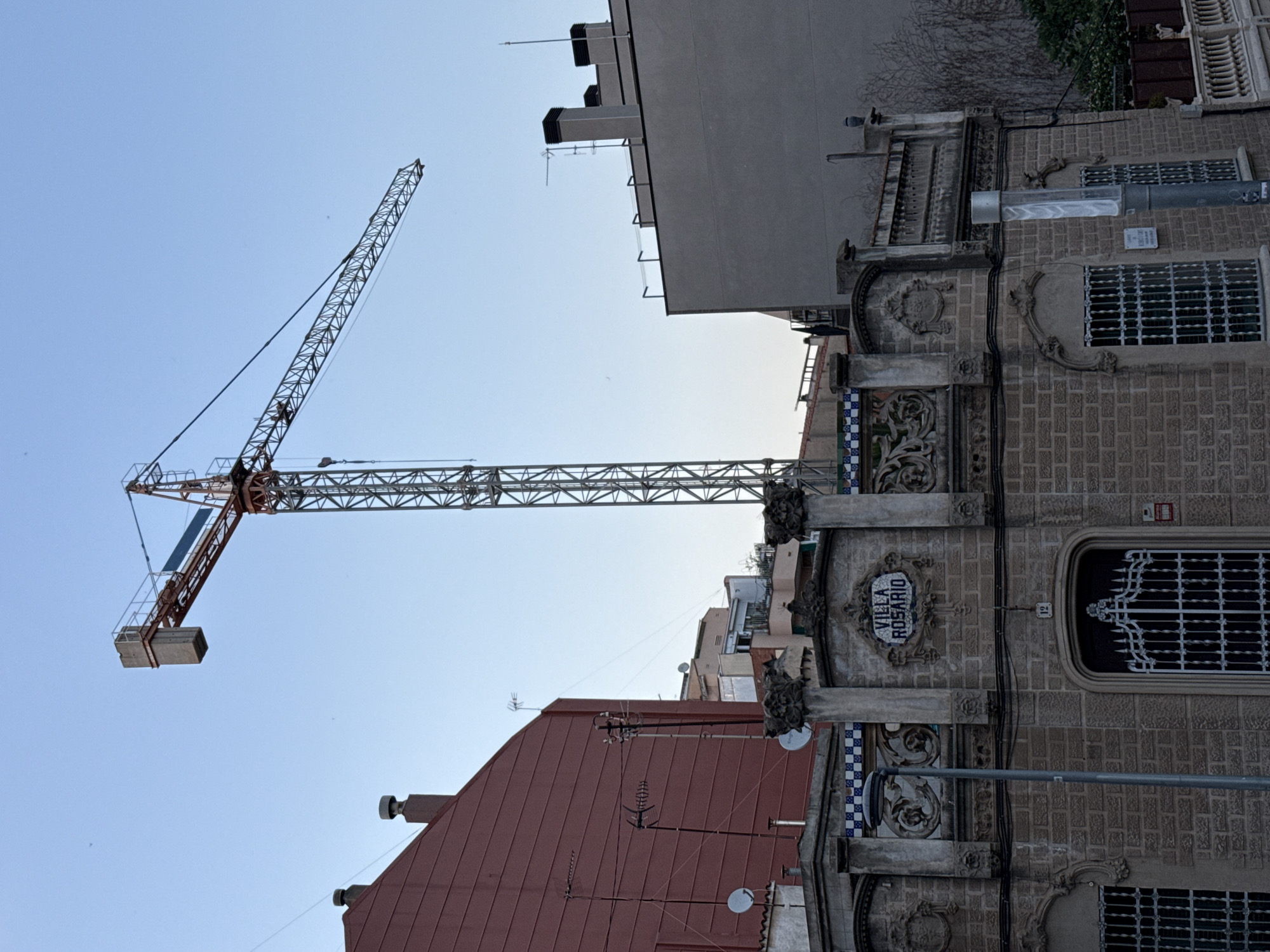 The image captures a street view in Barcelona, Spain, focusing on the facade of a building with the sign "VILLA ROSARIO" above its entrance. The building, constructed with light-colored brickwork, features ornate architectural details including decorative stone carvings, balustrades, and patterned tile accents along the roofline. A prominent street lamp stands to the left of the entrance. Above and behind this building, a large construction crane dominates the skyline, indicating ongoing development in the area. Adjacent structures, including a red building to the left and a gray building to the right, are also visible, along with various antennas and satellite dishes on the rooftops. The sky is clear and blue.