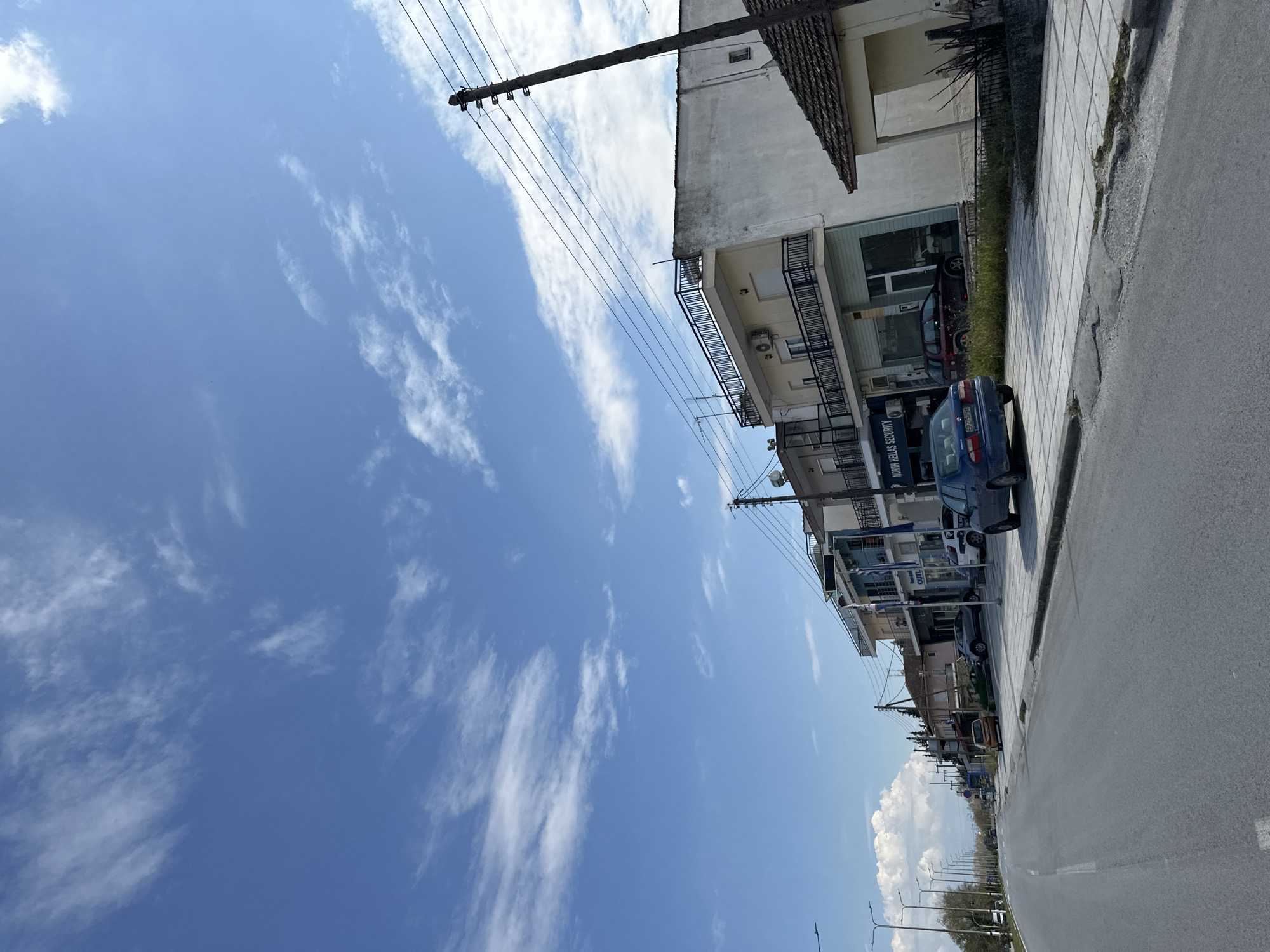 The image depicts a street scene in Lefkonas, Greece on a sunny day. The sky is a bright blue with wispy white clouds. Buildings line the right side of the frame, appearing to be two to three stories tall with balconies and air conditioning units. A dark blue car is parked along the sidewalk with a business advertisement on its side. Other parked cars are visible further down the street. Telephone poles and wires are strung above the buildings. The road takes up a significant portion of the lower right frame and appears to be asphalt. Streetlights are lined up along the street, further down. There are no people visible in the image.
