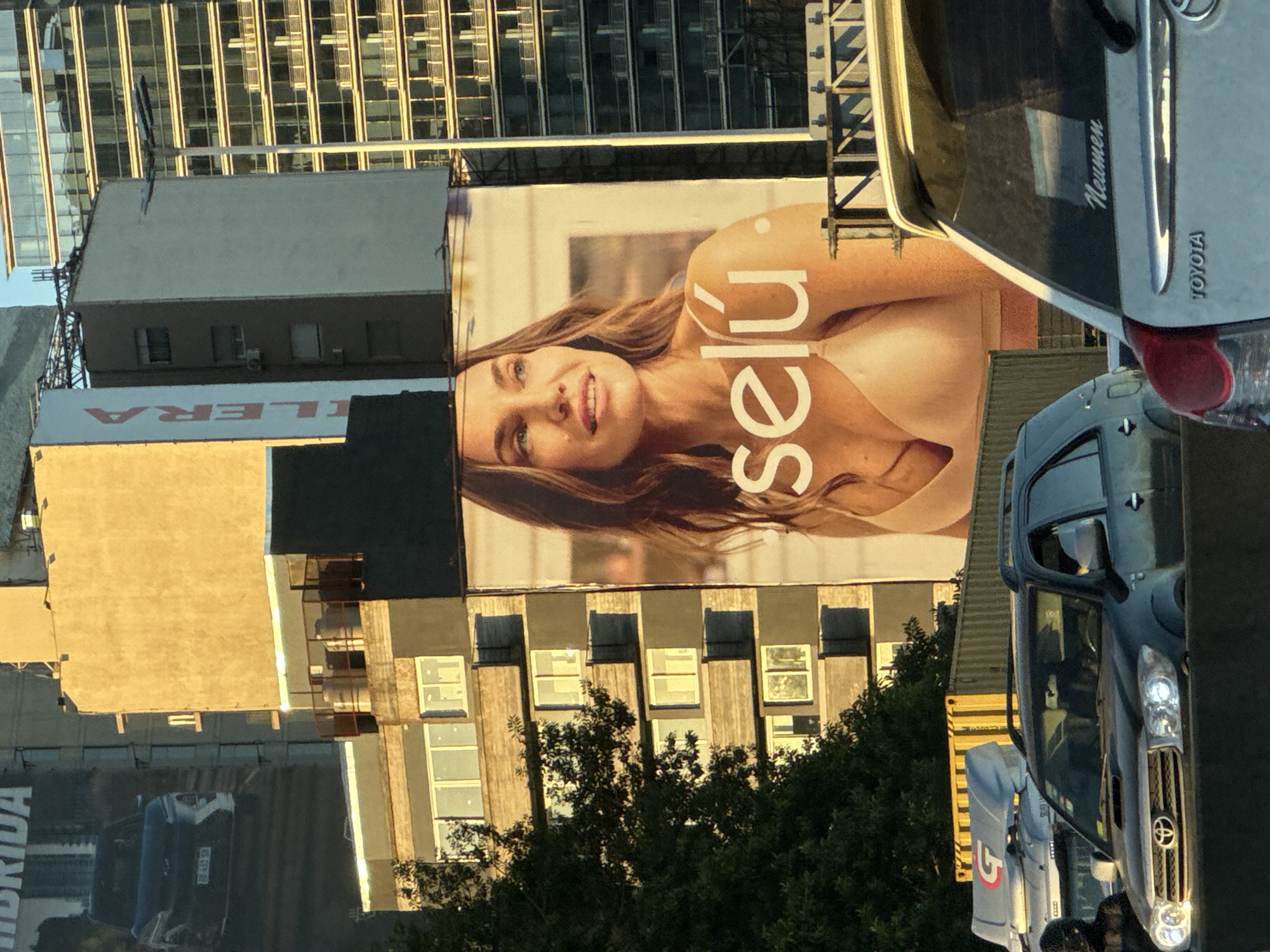 The image depicts a street scene in Buenos Aires, Argentina. A large billboard advertisement for "Selú" lingerie is prominently displayed on a building. A Toyota truck with "Naumen" branding is parked near the billboard. Other vehicles and buildings are visible in the background, including a modern glass skyscraper. Trees line the street, and people can be seen walking on the sidewalk. The overall impression is of a typical urban environment in Buenos Aires.