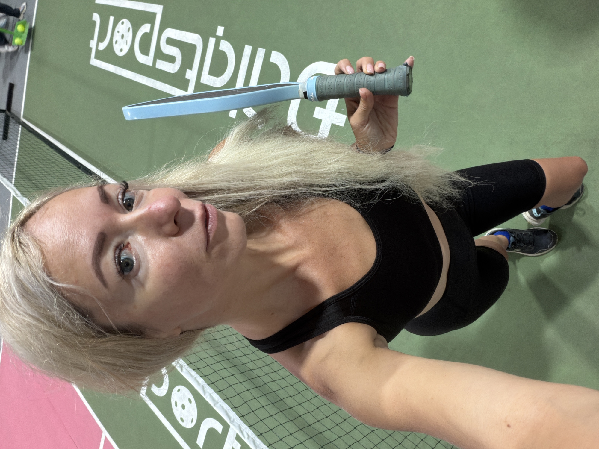 The image shows a blonde woman holding a pickleball paddle on a pickleball court. The court is green with white lines and a logo that says "Clipsport". A net is visible in the background. The woman is wearing a black sports bra and black shorts. She is looking at the camera and smiling. The location is a pickleball court in an indoor facility. There is no indication that this location is in Da Lat, Vietnam.