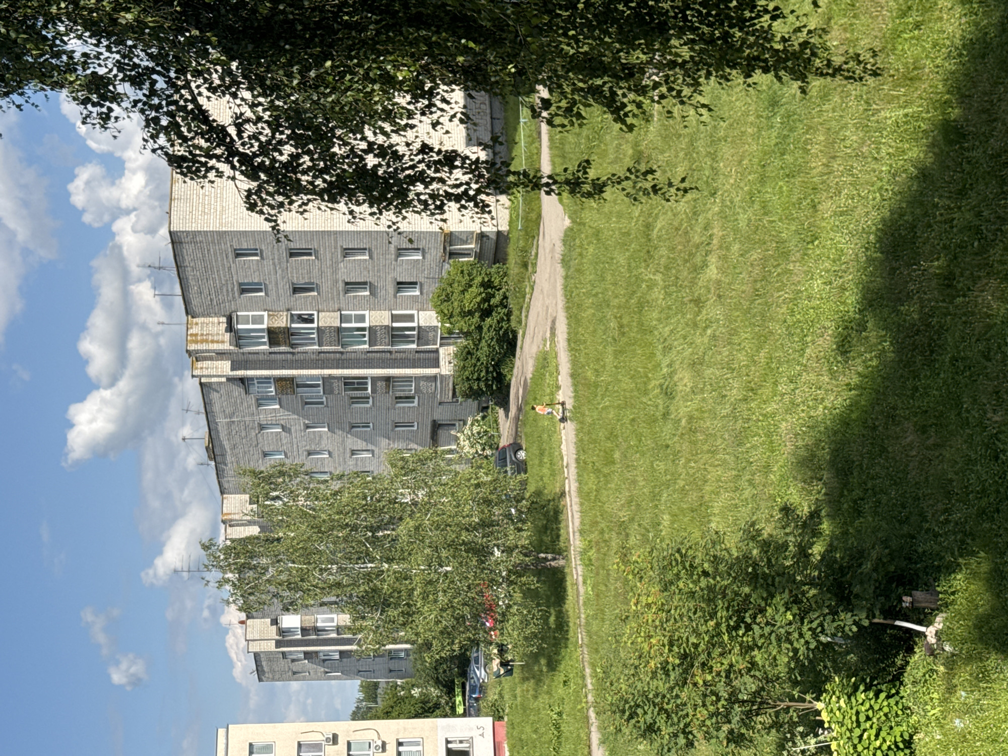 The image depicts a residential area in the Яромінскі сельскі Савет, Belarus, on a sunny day with scattered clouds. In the background, a multi-story apartment building constructed of light-colored bricks is visible. The building has numerous windows and balconies. In front of the building, a line of parked cars is seen. A young person is riding an electric scooter on a paved path in the middle ground. Lush green grass covers the foreground and extends towards the buildings. A large birch tree with green leaves stands prominently in the mid-ground, and the branches of another tree with darker leaves partially obscure the right side of the image. The overall scene conveys a typical summer day in a Belarusian residential neighborhood.