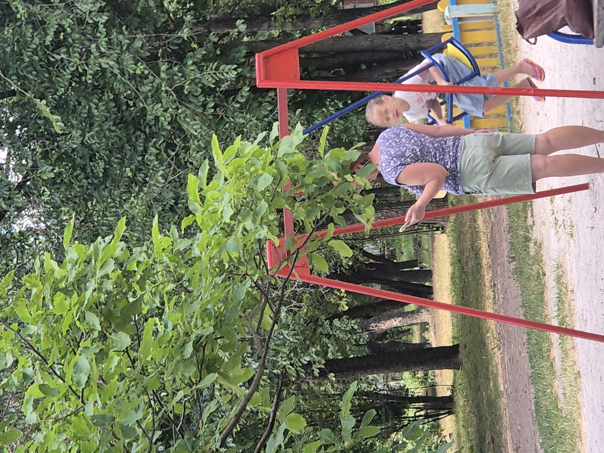 In Sozonivka, Ukraine, a woman in a purple patterned shirt and light green shorts stands to the left, looking at her phone. To her right, a young girl in a white t-shirt and blue shorts is on a swing set with a red frame and yellow and blue seat. The swing set is in a park-like setting with trees and grass. A brown bag is visible to the right of the swing set.