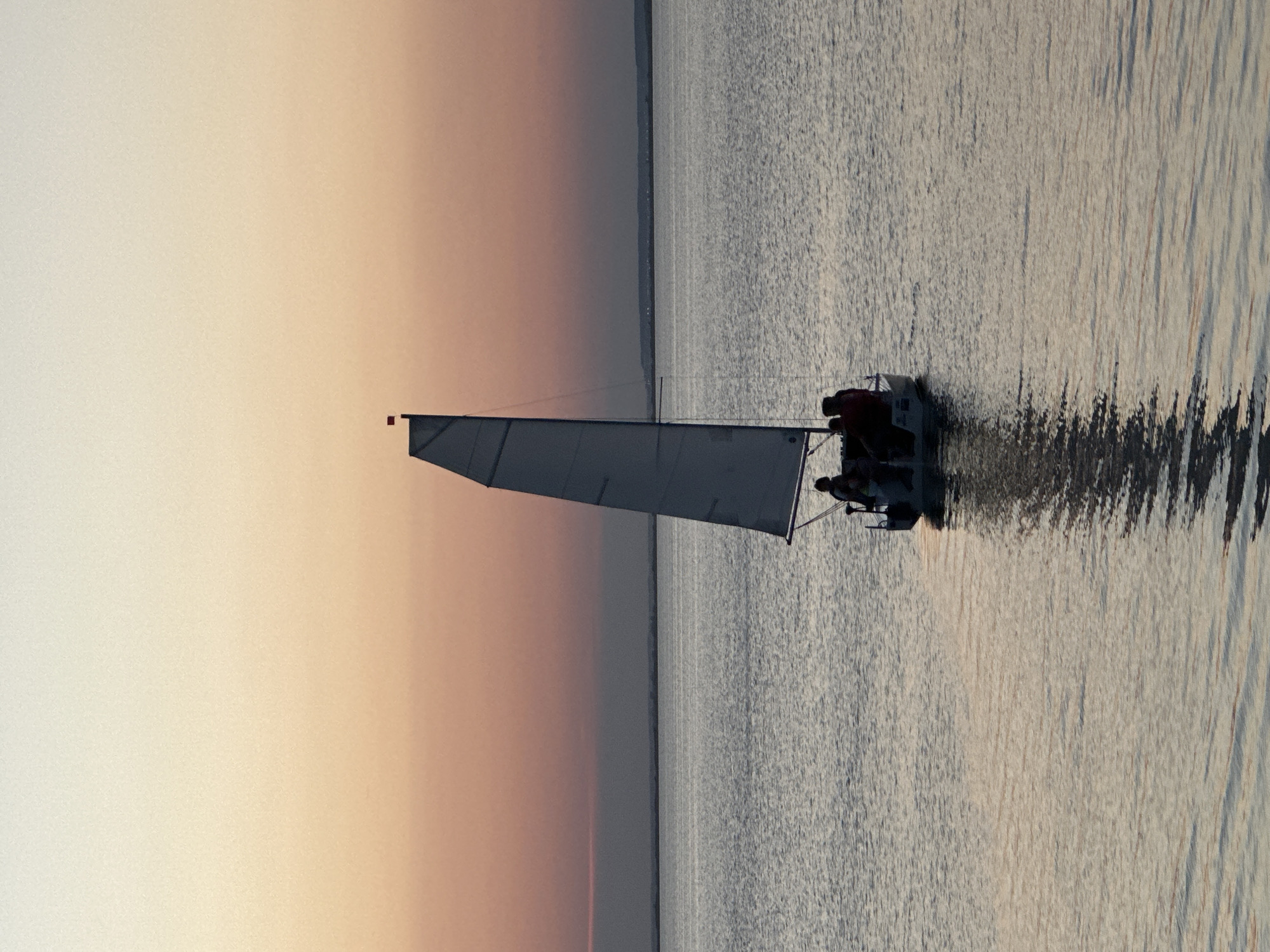 The image depicts a sailboat with several people on board sailing on a body of water. The sky above is a gradient of colors, transitioning from a pale yellow at the top to a peachy orange and then a muted pink closer to the horizon. The water is calm with gentle ripples and reflects the colors of the sky. In the distance, a faint landmass is visible on the horizon. The sailboat has a white sail and a dark hull. The people on the boat are silhouetted against the sky, making their features indistinguishable. The overall scene evokes a sense of tranquility and a serene evening on the water. This scene is likely a coastal area or a large lake.