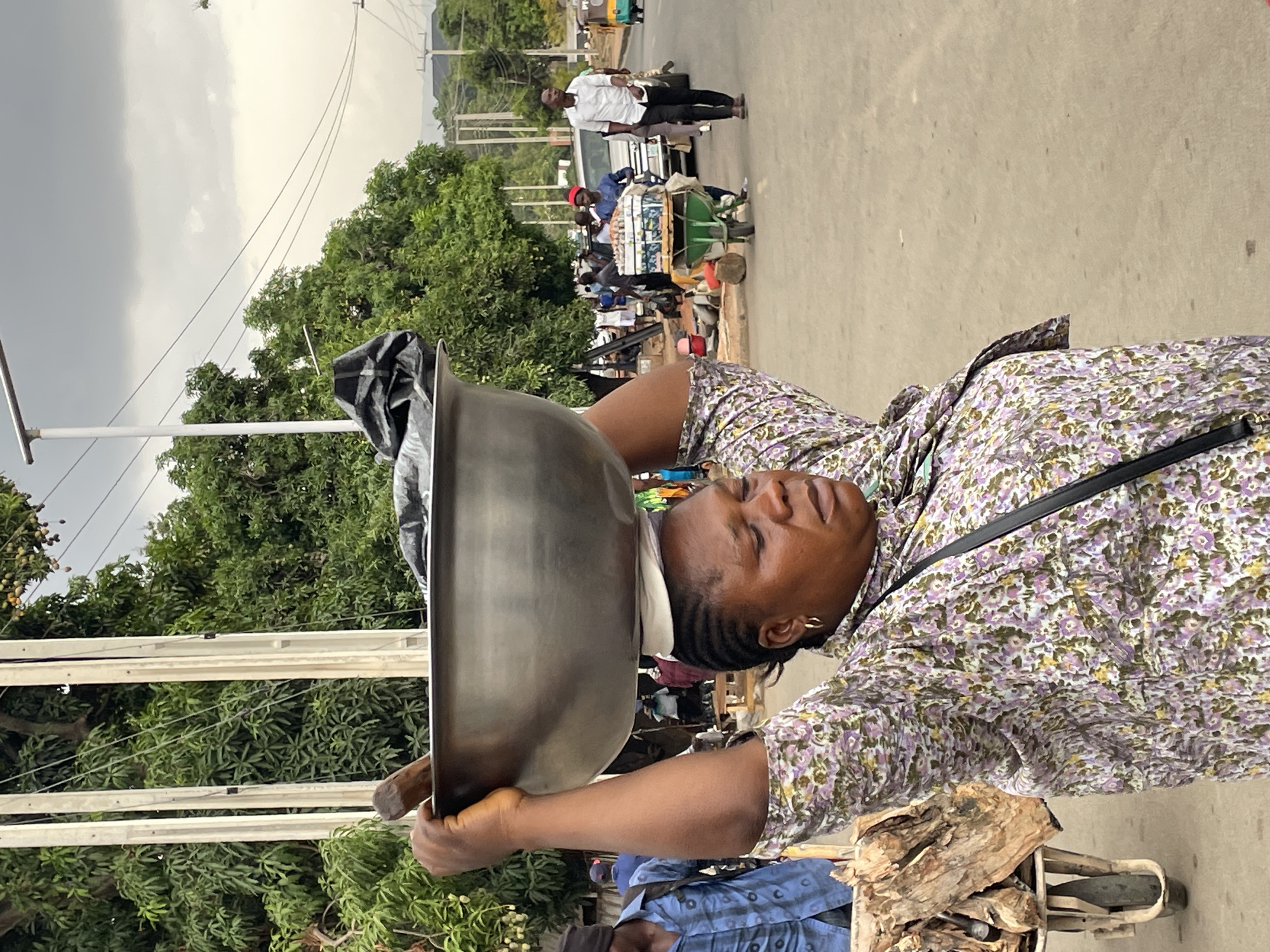 The image captures a vibrant street scene in Jos, Nigeria, under an overcast sky. The main subject is a woman in the foreground, positioned in the lower right portion of the frame (due to the image's rotation). She is carrying a large, grey metallic bowl on her head, supported by a white cloth. Her hands are also placed on the bowl, helping to steady it. She has dark skin, braided hair, and wears a light-colored top with a distinctive purple, green, and yellow floral pattern, along with a black strap across her shoulder. Her eyes are closed or downcast, giving her a serene expression.
Behind her, the street is busy with people and activity. A man in a white shirt and dark trousers walks further down the road. Behind him, a white car is visible. A street vendor's cart, laden with numerous small, packaged goods, is prominent in the mid-ground, with a person in a red hat standing nearby. Other individuals are scattered throughout the background, suggesting a lively environment.
Lush green trees line the street, indicating an area with significant vegetation. Utility poles and overhead wires crisscross the upper portion of the frame against the cloudy, light grey sky. In the bottom right corner, a wooden cart or stall is partially visible, filled with what appears to be firewood or rough-cut wood pieces. Another person, wearing a blue patterned shirt, is partially seen behind this cart. The image depicts daily life, showcasing typical street vending and head-carrying practices in the region. There is no clearly legible text visible in the frame.