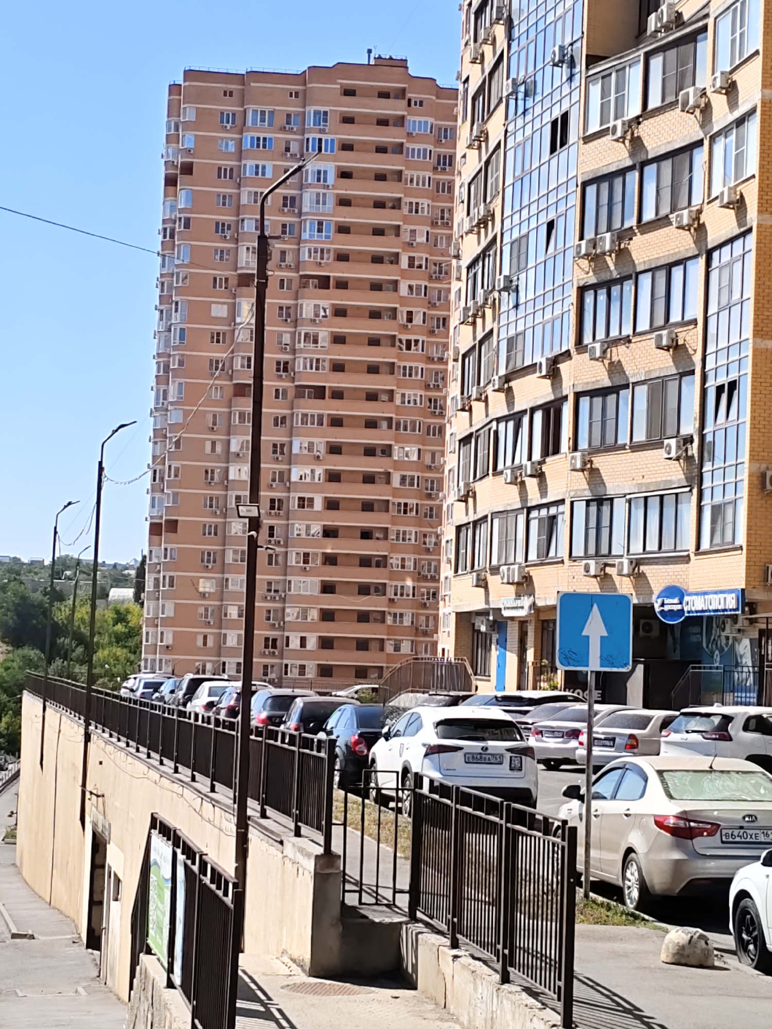 The image captures a street scene in Rostov-on-Don, Russia, with a focus on modern apartment buildings and parked cars. The foreground features a raised walkway with a dark metal railing, bordered by a beige concrete wall on the left. Several streetlights with a distinctive shape are visible along the walkway.
To the right of the walkway, a row of parked cars is lined up along the street. These include a white Lexus with license plate E868DA751, a light-colored Kia with license plate B640XE161, and other vehicles. A blue traffic sign with a white upward-pointing arrow is mounted on a pole in front of a brick building. This building, with its yellow brick facade and numerous windows, appears to be a commercial establishment, as evidenced by a circular blue sign with text in Russian that reads "СТОМАТОЛОГИЯ" (Dentistry).
In the background, two tall apartment buildings dominate the skyline. The building on the left is a slender, peach-colored structure with many small, uniform windows and balconies. The building on the right is wider, with a modern design featuring a yellow brick facade and large, reflective glass windows that create a mosaic-like effect. Air conditioning units are visible mounted on the exterior walls of this building.
The sky is clear and blue, indicating a sunny day. Green trees and vegetation are visible on the left side of the image, suggesting the presence of a park or natural landscape beyond the urban development. The overall impression is one of a contemporary urban environment.