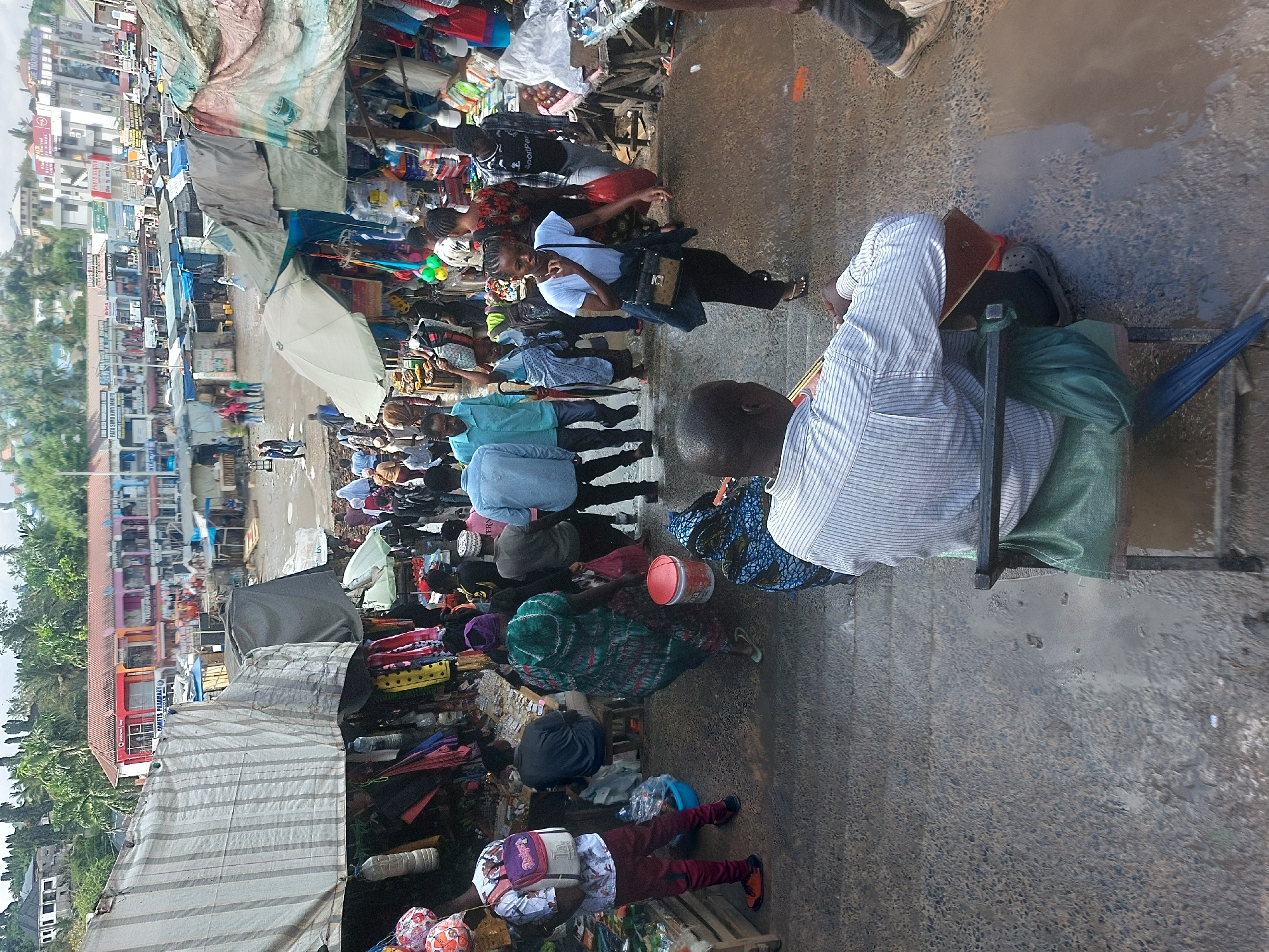 This image captures a vibrant and crowded street market scene in what appears to be Dar es-Salaam, Tanzania, under bright daylight, possibly after some rain given the puddles on the ground.
**Scene and Setting:** The street is bustling with activity, featuring numerous makeshift market stalls constructed from various tarpaulins (grey, blue, striped) and fabrics. These stalls line one side of a paved thoroughfare, while multi-story buildings with shops on their lower levels form the backdrop on the other side. The overall atmosphere is busy and commercial.
**Subjects - People:** Many people are present, indicating a high level of activity. In the right foreground, a man in a light-colored striped shirt is seated on a simple chair, facing left, observing the scene. To his left, a person in a light blue top is partially visible. Further into the scene, a woman in a white t-shirt, dark pants, and holding a handbag is walking towards the viewer, offering a slight smile. Other individuals are seen walking, browsing, or attending to their stalls, dressed in a variety of casual attire, some with head coverings. A person wearing maroon pants and a white t-shirt with a backpack walks away from the viewer in the middle ground.
**Subjects - Objects:** The market stalls display a diverse range of goods, including clothing, plastic items, bottles, and other general merchandise. Several open umbrellas, predominantly white, are visible, likely providing shade from the sun or protection from light rain. Various containers, such as a red bucket, are scattered among the stalls. The ground is a mix of paved surface and visible puddles of standing water.
**Activity:** The primary activity is that of a lively street market where vendors sell goods and people shop, walk through, and socialize. It's a typical scene of daily commerce and urban life.
**Notable Details:** The light suggests it's daytime. The puddles indicate recent rain or poor drainage. Visible text on signs in the background includes "SADAKA," "PALTRY," and "SPORTS PHARMACY," offering small clues about the businesses operating there. The scene conveys a sense of everyday industriousness and community.