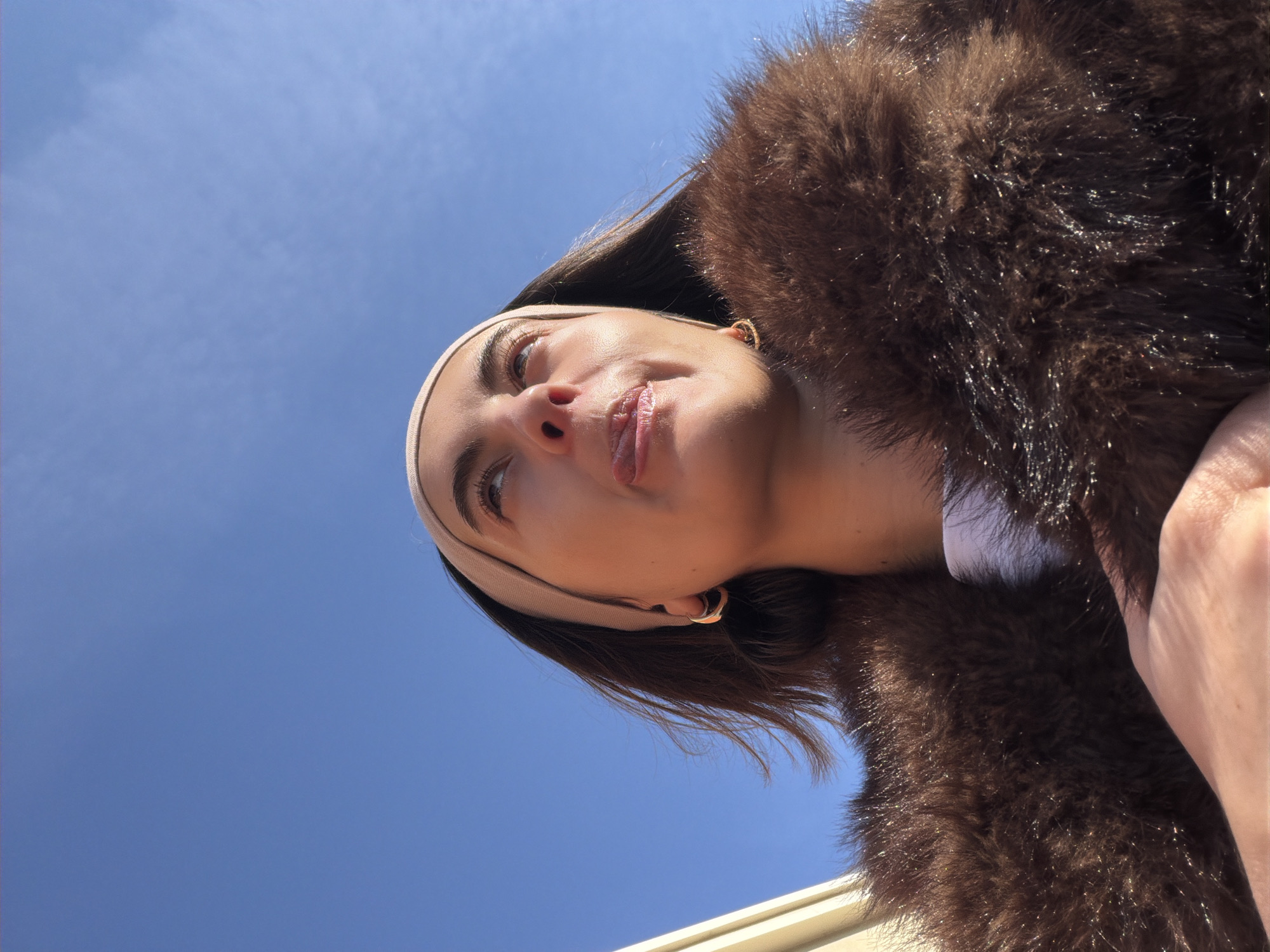 A low-angle shot depicts a woman wearing a brown fur coat and a neutral-colored headband. She is positioned against a clear blue sky with faint wisps of clouds. The angle suggests the photo was taken from below, looking up at the woman. Her face is in focus, revealing details such as her makeup, facial features, and a slight smirk. Her hair is a dark brown color, partially visible beneath the headband and near her ears. Her gold earrings are visible. A portion of the top of a building is barely visible at the bottom of the frame. There are no specific landmarks or contextual elements to determine the location is in Mascouche, Canada.