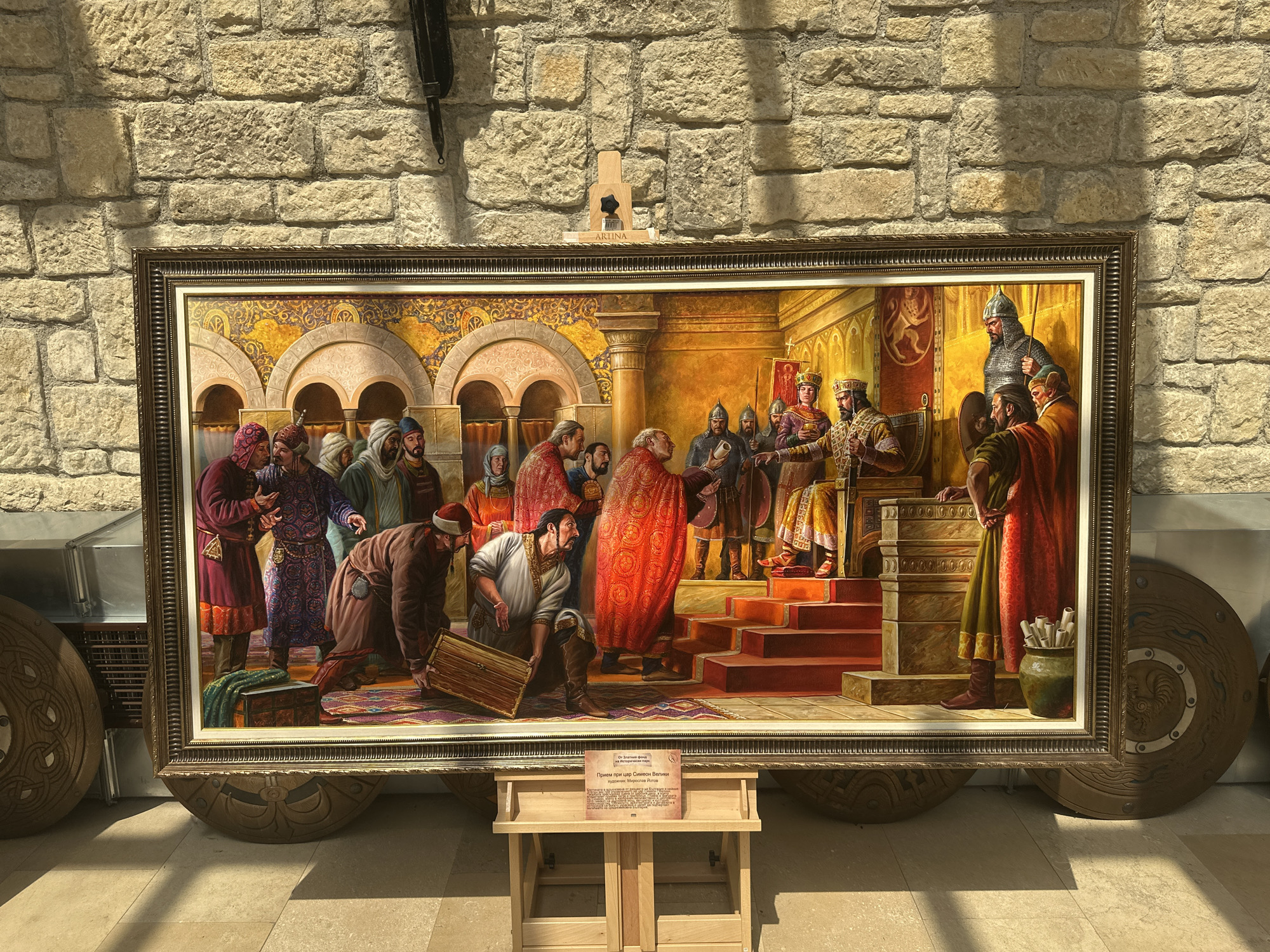 The image shows a large framed oil painting depicting a historical scene of a royal audience. The painting is displayed on a light-colored tiled floor against a textured stone wall. In the foreground, a wooden easel holds a sign with text in Cyrillic. The painting itself shows a group of figures dressed in historical attire within an opulent interior. The central figures appear to be a king or ruler seated on a throne or raised platform, with other individuals presenting offerings or engaging in conversation. The figures wear elaborate robes and headwear, suggesting a setting in the past, possibly the Middle Ages. The interior features ornate architecture with arches and patterned walls. To the left and right of the painting, large circular shields with intricate designs are visible, leaning against the wall. The lighting in the scene suggests indoor illumination with some natural light casting shadows on the floor and wall. The location is identified as Neofit Rilski, Bulgaria, implying this is likely a museum or historical exhibition space.