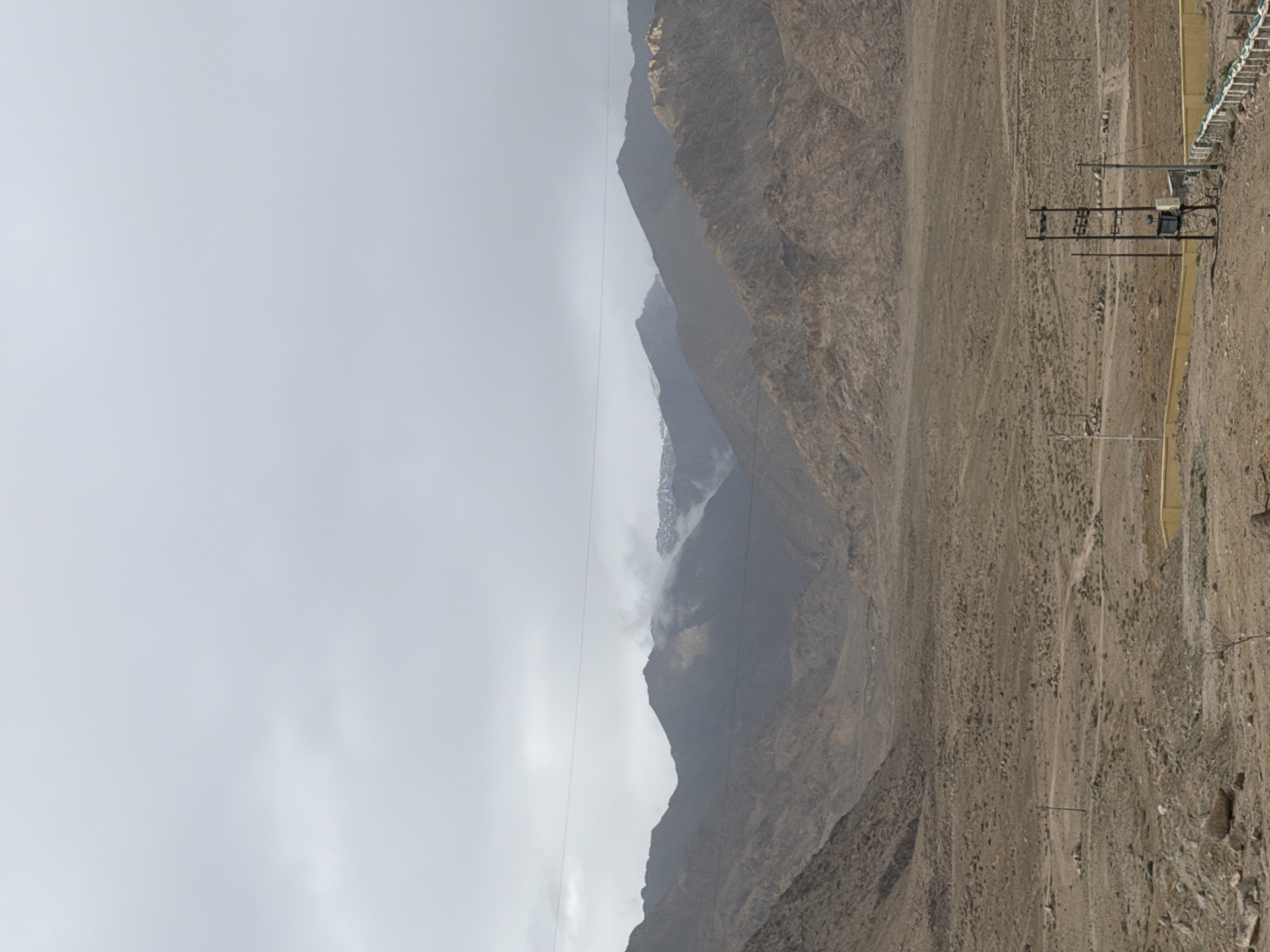 This is a landscape photograph taken in Chumkar, India, under a cloudy sky. The image captures a vast, arid valley with rugged, barren mountains in the background. The mountains are layered in shades of brown and grey, with some peaks appearing snow-capped. In the foreground, the ground is dry and dusty, scattered with rocks and sparse vegetation. A yellow-walled enclosure is visible in the lower right portion of the frame, next to a metal utility pole with electrical equipment. Power lines are strung across the sky, bisecting the view of the mountains. There are no people or animals visible in the image. The overall scene depicts a remote, undeveloped, and harsh environment.