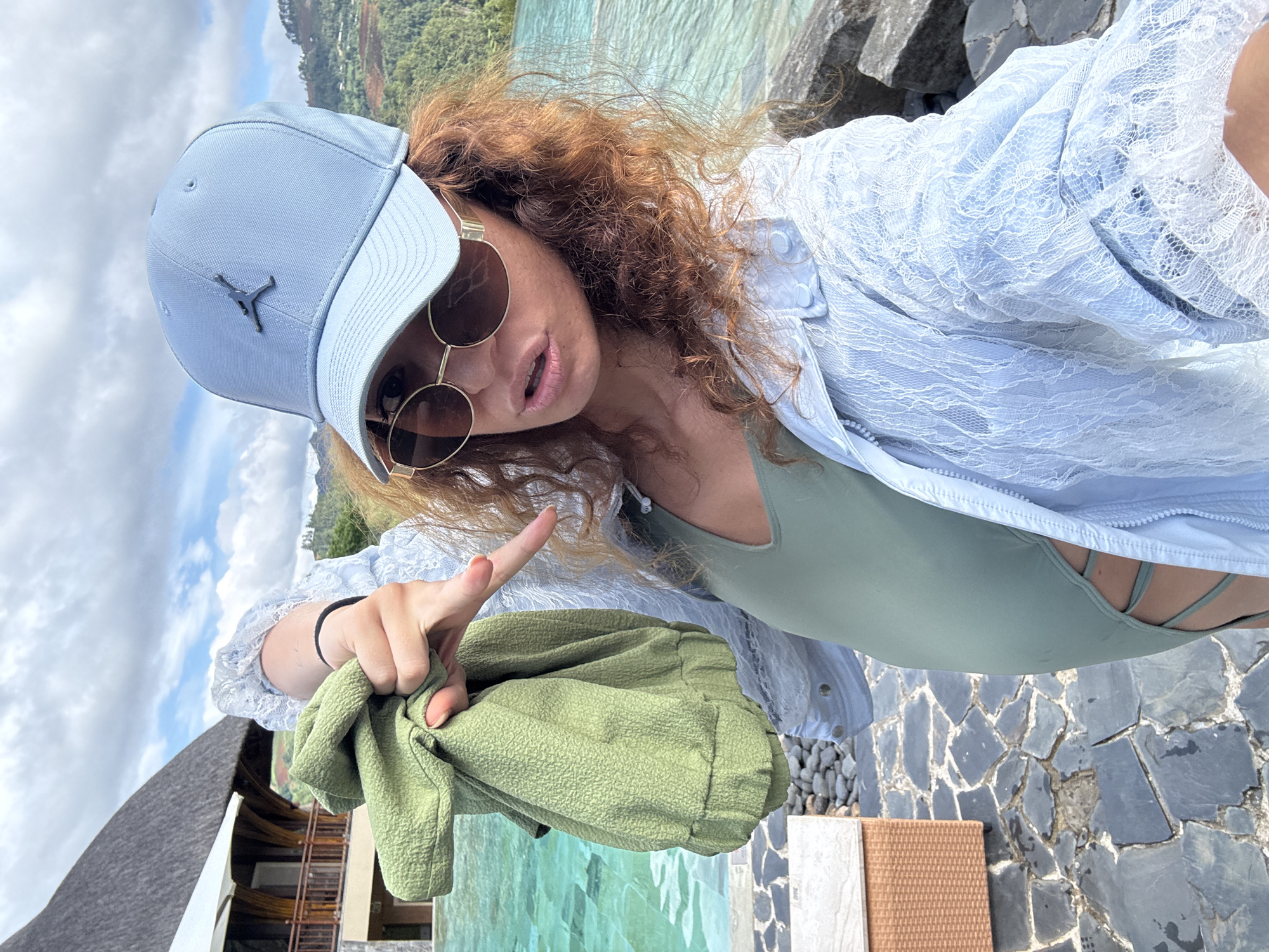 A selfie-style outdoor shot, angled diagonally, captures a person, likely female, engaged in a vibrant setting. The subject has curly, reddish-brown hair and wears a light blue baseball cap featuring a dark Jordan 'Jumpman' logo on the front. Round, gold-rimmed sunglasses obscure her eyes, and her lips are slightly parted, suggesting an active expression. She is dressed in a light blue long-sleeved lace cover-up or shirt worn over a sage green top, possibly a swimsuit, which reveals intricate strap detailing at the back. With her right hand, she points directly forward or slightly towards the viewer, while her left hand holds a piece of textured, olive-green fabric.
The background presents a picturesque landscape: a body of clear, light blue-green water, indicative of an ocean or large lake, extends to the horizon, meeting a backdrop of lush, green forested mountains and hills. The sky above is bright with scattered white clouds, suggesting a sunny day. In the lower left, part of a building with a dark, sloped roof and visible wooden railings can be seen. The foreground consists of an irregular mosaic of dark stone paving and some lighter, textured surfaces. The scene conveys a relaxed atmosphere in a scenic, possibly resort-like location, consistent with Púng Luông, Vietnam. The time of day appears to be bright daylight.