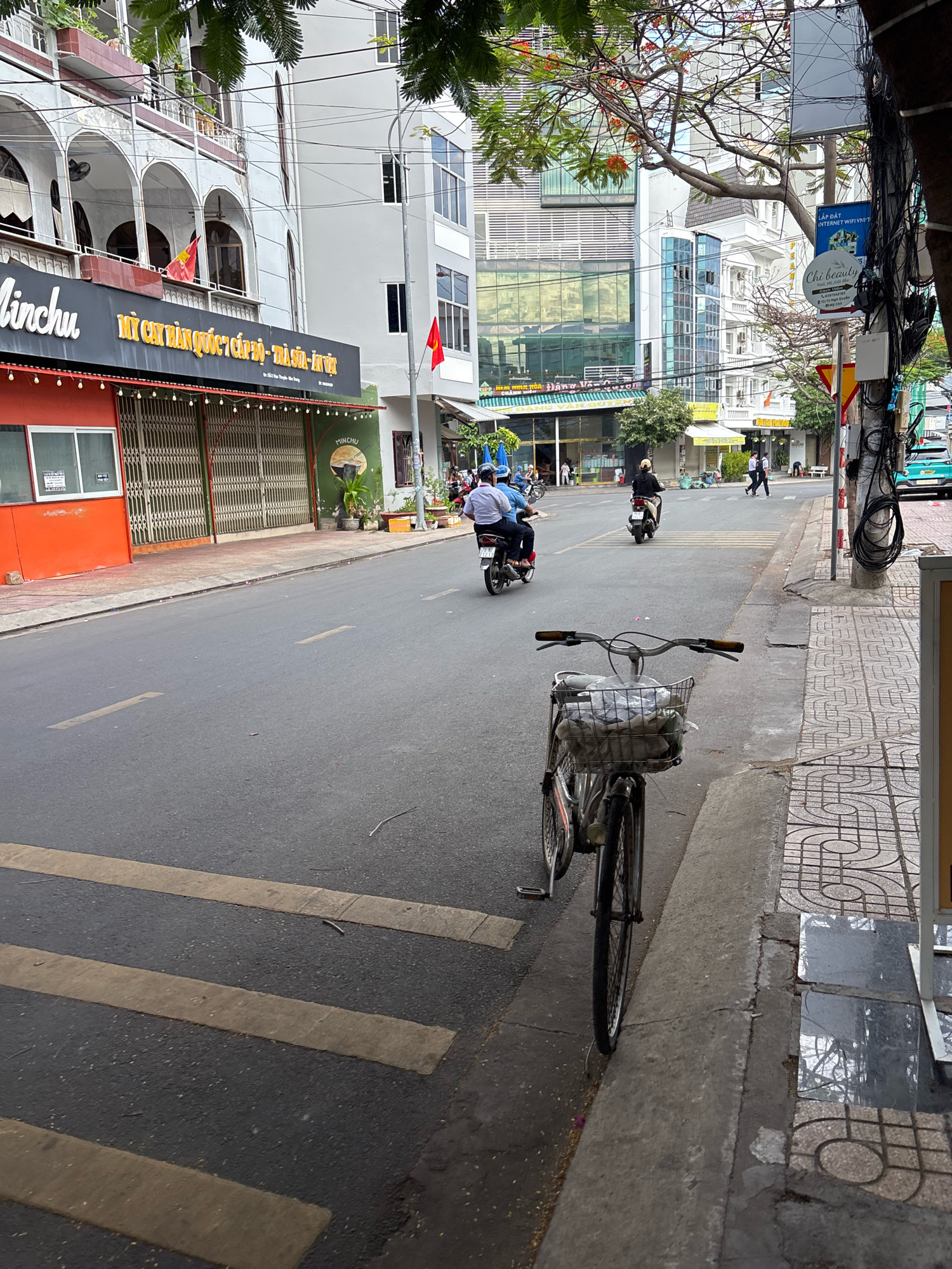 The image depicts a street scene in Nha Trang, Vietnam. In the foreground, a bicycle with a basket filled with items is parked on the sidewalk next to a crosswalk. The street is paved and appears wet in some areas. Several motorcycles are visible on the road, with people riding them. Buildings line the street, featuring a mix of architectural styles, including modern structures with glass facades and older buildings with arched windows and balconies. Vietnamese flags are displayed on some of the buildings. The environment suggests a bustling urban area with commercial activity, as indicated by the storefronts and signage. The time of day appears to be daytime, with natural light illuminating the scene.