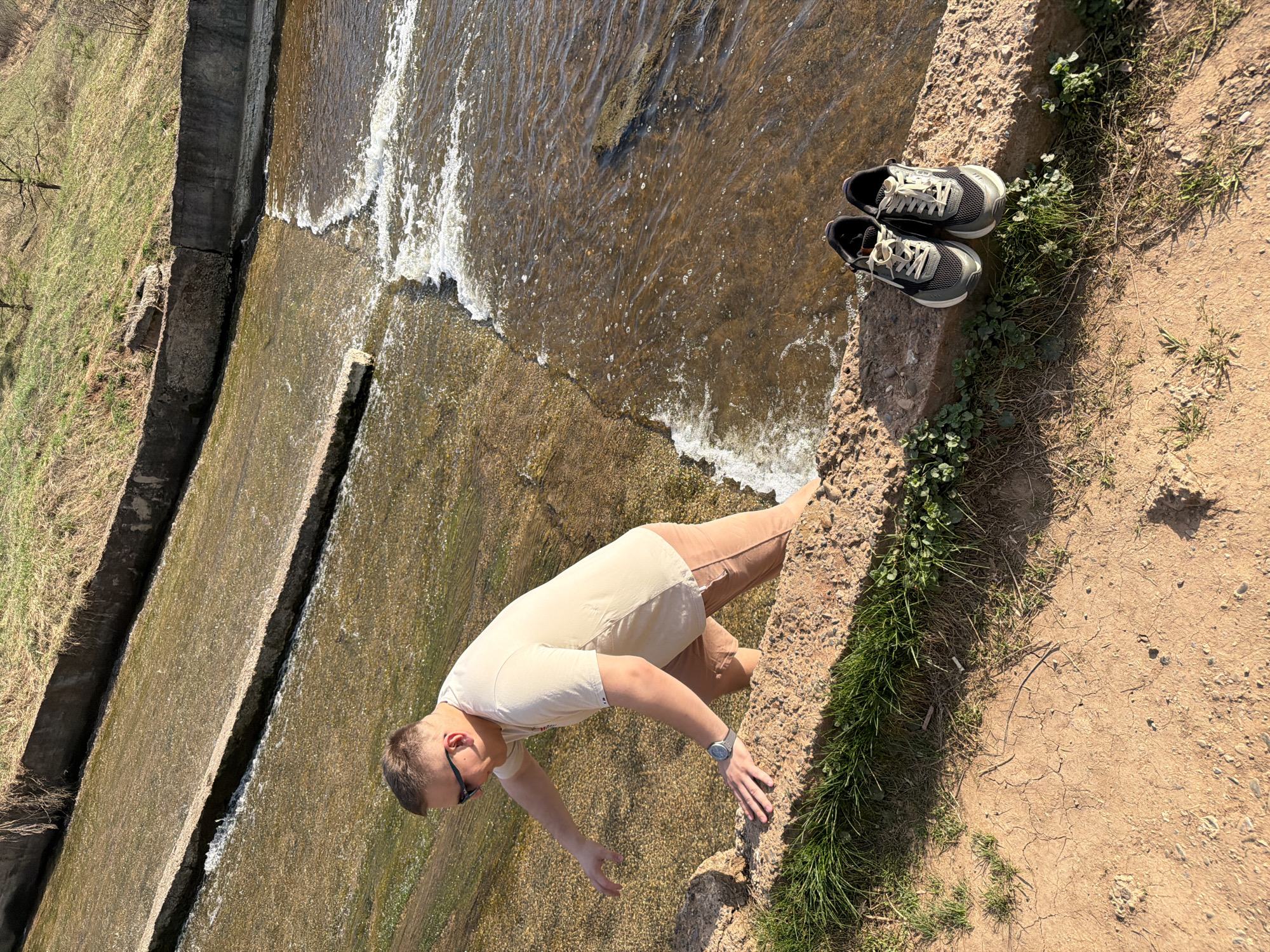 The image shows a man leaning over a stepped concrete waterfall structure in what appears to be a natural or park setting. The man is wearing a light-colored t-shirt, tan pants, and sunglasses. He is near a pair of sneakers. The ground is covered in dirt and grass. The location may be Kamennoye, Russia.