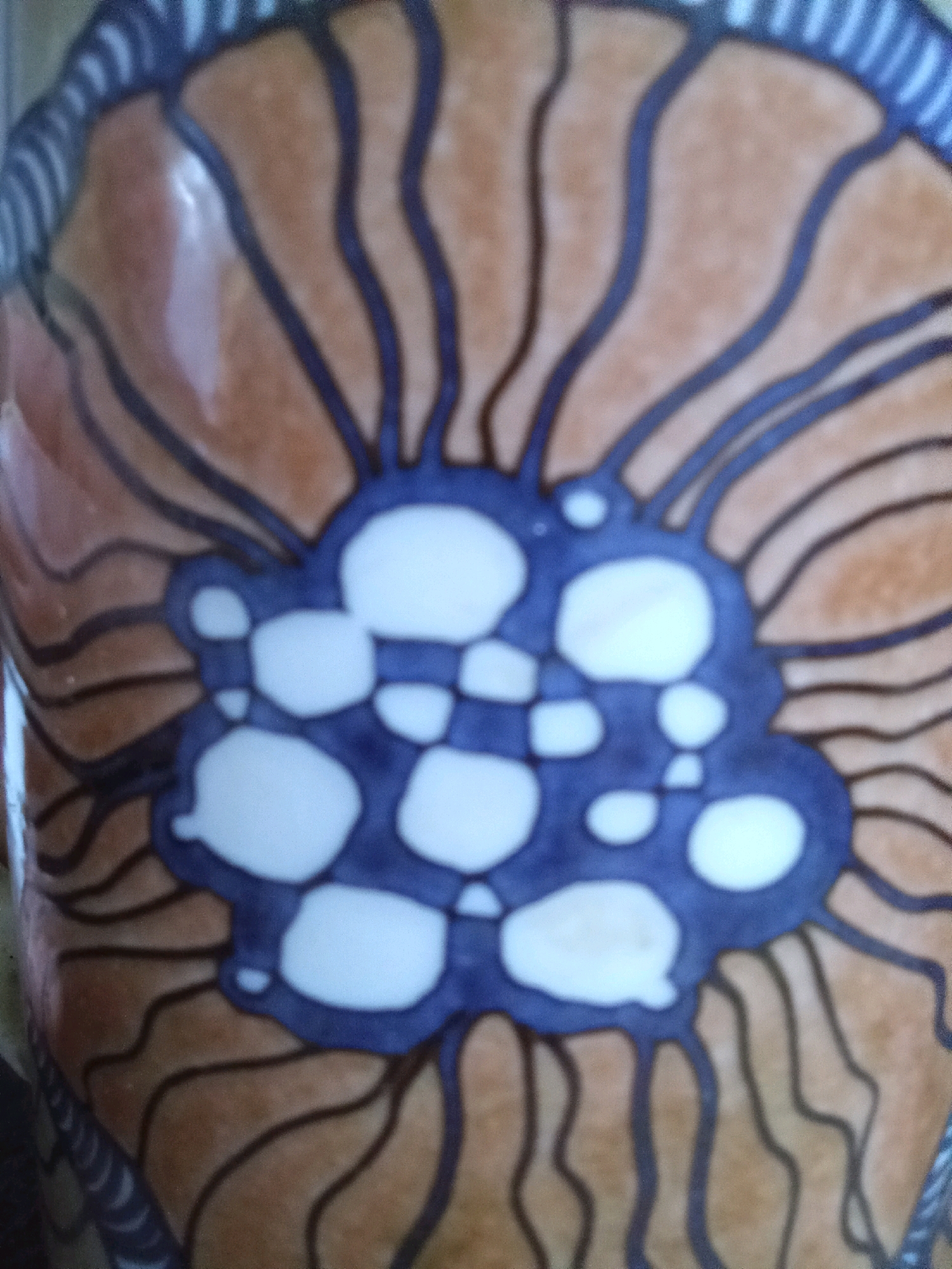The image shows a close-up view of a ceramic object, likely a vase or pot, with a decorative pattern. The pattern consists of brown and orange wavy lines radiating outwards from a central blue and white floral or abstract motif. The blue and white design is composed of irregularly shaped white patches outlined in blue, resembling petals or cells. The background of the ceramic is a warm, glossy brown-orange color. The top edge of the object has a border with alternating blue and white wavy lines. The image is slightly out of focus, with reflections visible on the glossy surface of the ceramic. The specific location in Aktau, Kazakhstan, cannot be determined from the image alone.