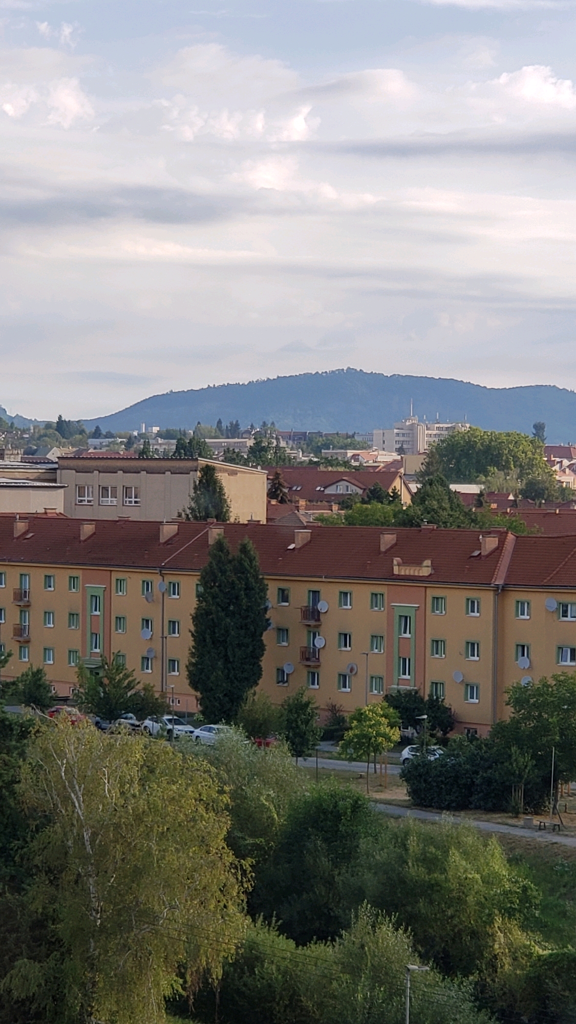 The image shows a view of a residential building in the city of Prešov, Slovakia. The building is a multi-story apartment complex with a yellow facade and a reddish-brown tiled roof. It has many windows and balconies, some of which have satellite dishes attached. In front of the building, there are trees and a road with a few cars parked. In the background, there are other buildings and a large hill covered in trees. The sky is cloudy. The scene appears to be a typical urban landscape.