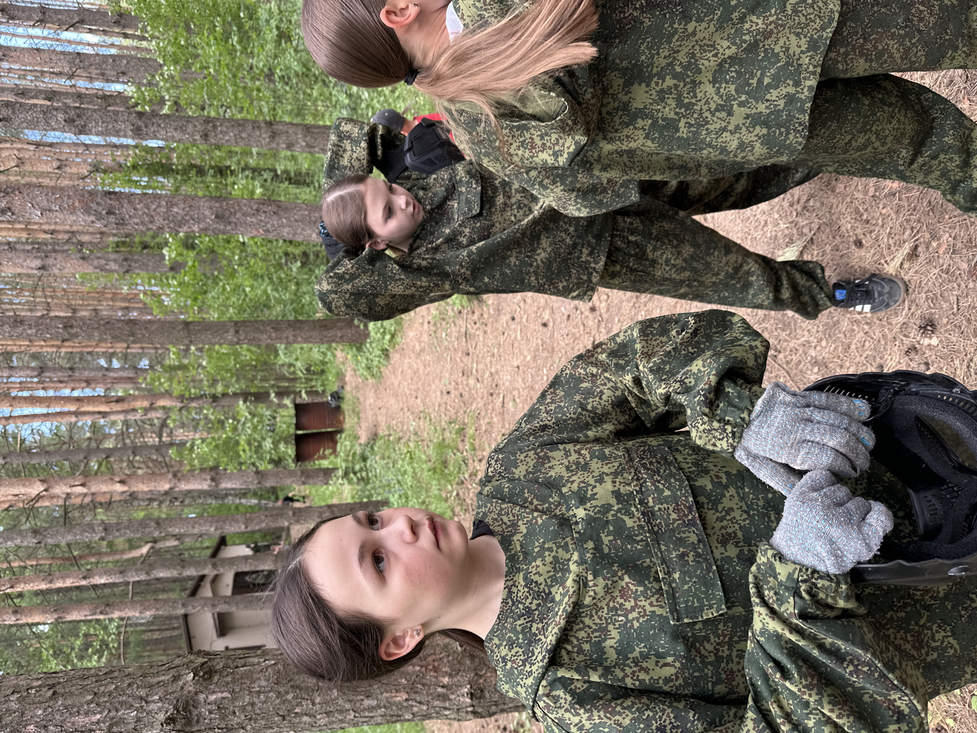 The photograph captures three individuals in a wooded area, possibly near Votkinsk, Russia, based on the presence of Russian military-style camouflage uniforms. The location is characterized by tall pine trees and a ground covered in pine needles and dirt. A two-toned structure is partially visible in the background.
A young woman in the foreground wears a camouflage uniform and grey work gloves, holding a black helmet. She is looking upward and to the left. In the middle ground, another individual, also in camouflage, has their hands behind their head. A third person, primarily visible from the back, stands behind and to the right of the second individual, also in a camouflage uniform.