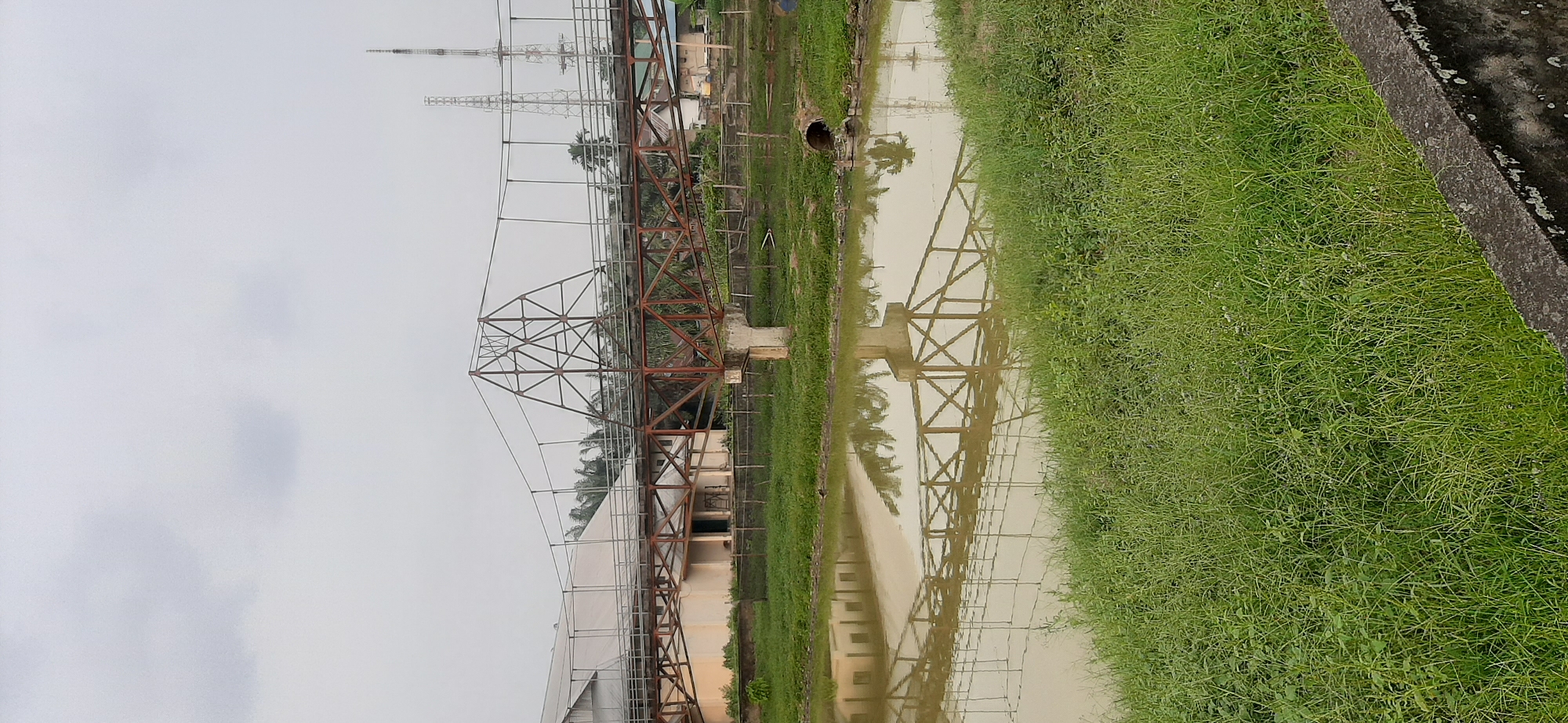 This is an outdoor, vertical, eye-level shot of a suspension bridge over a body of water, likely a river or canal, in the city of Jambi, Indonesia. The bridge, made of weathered metal and concrete supports, features a central truss structure and suspension cables. On the far side, a long, light-colored building with a sloped roof is visible, accompanied by palm trees and other vegetation. In the background, two tall communication towers pierce the overcast sky. The water reflects the bridge and surrounding greenery, with a concrete embankment and lush grass in the foreground. A large drainage pipe exits the embankment into the water on the right side of the frame. The scene appears quiet and undeveloped, with no people or vehicles present.