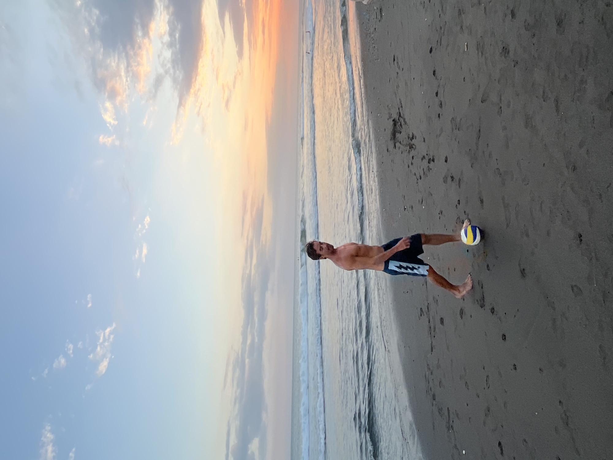 A shirtless man wearing dark blue and white patterned swim trunks is walking on a sandy beach, kicking a volleyball. The sun is setting, casting a warm orange and pink glow across the sky and reflecting on the ocean waves. The beach is dark gray sand with footprints. In the distance, several people can be seen in the water. The scene is located in North Kuta, Indonesia.