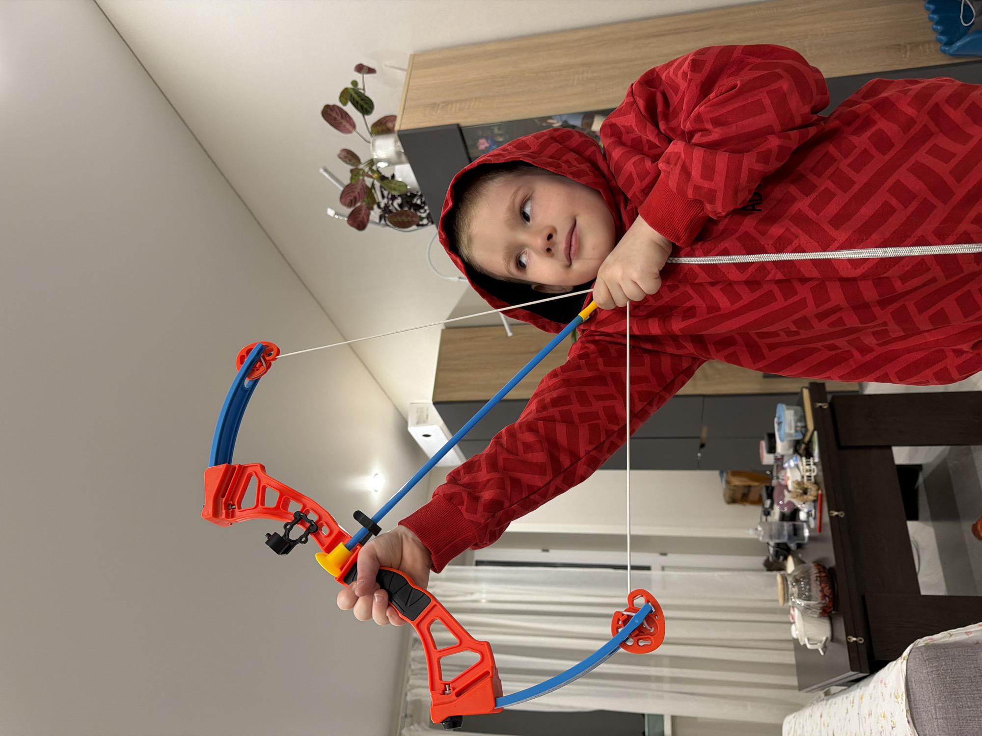 A young child in a red hooded sweatshirt is holding a toy bow and arrow inside a home in Засечное, Russia. The bow is primarily orange and blue with a white string. A table cluttered with various items is visible in the background.