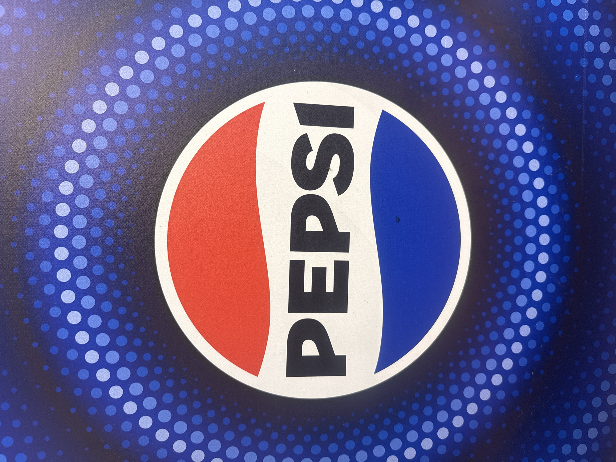 The image is a close-up, eye-level shot of a Pepsi logo against a blue, dotted background. The Pepsi logo is circular, with a red half-circle at the top, a blue half-circle at the bottom, and a white wave-like band separating them. The word "PEPSI" is prominently displayed in bold, black, sans-serif font across the white band. The background consists of concentric circles of dots, primarily in shades of blue, that become smaller and darker towards the edges of the frame. There are no people or specific environmental details visible that would indicate a location within a city.