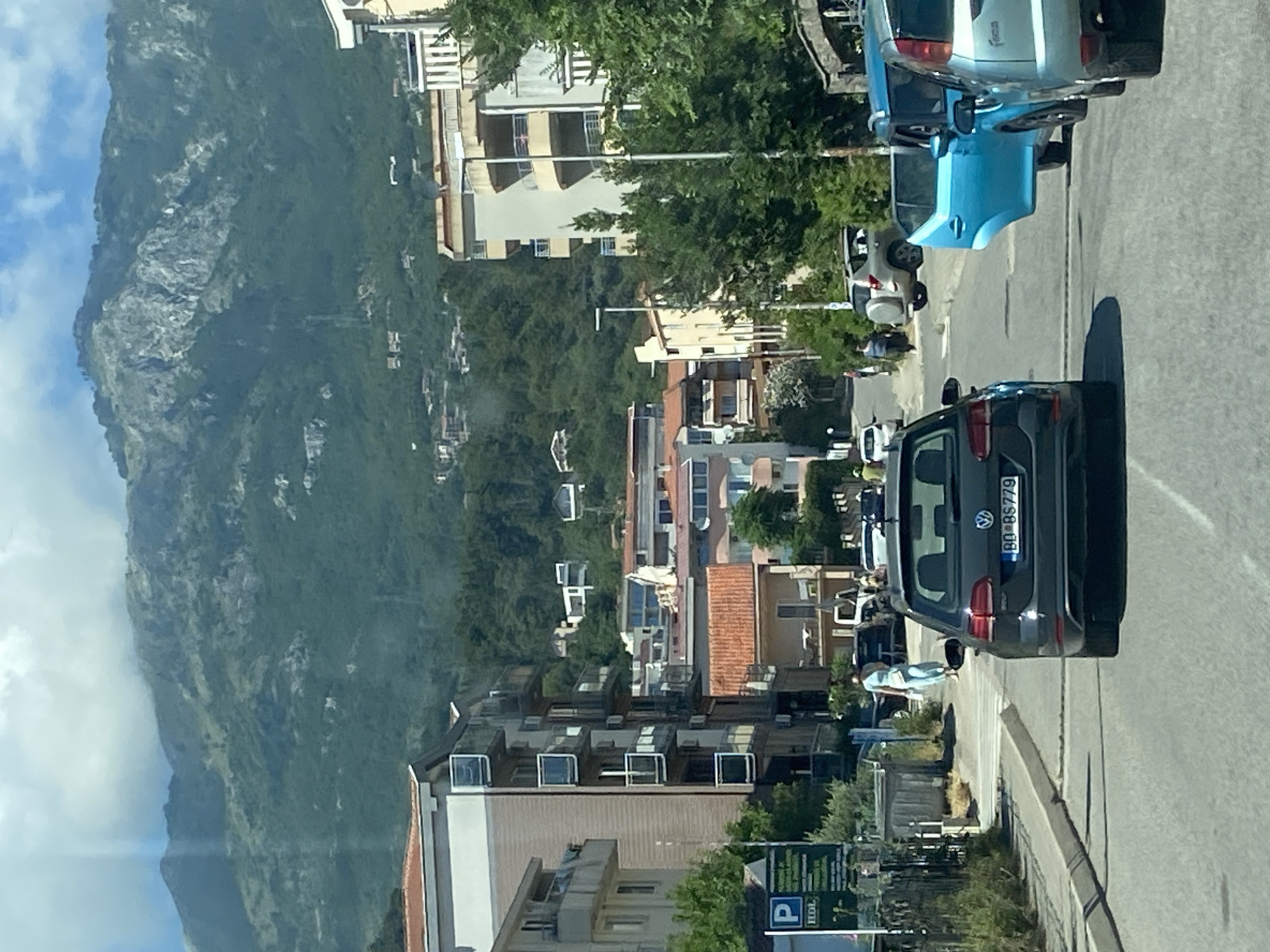 The image depicts a street scene in Budva, Montenegro, characterized by apartment buildings and a mountainous backdrop. In the foreground, a dark-colored Volkswagen Golf with license plate "BO BS779" is prominently featured, indicating it is the primary subject of the photograph. To its right, a light blue car with its passenger door open is visible, suggesting a recent stop or activity. Further down the street, other vehicles and pedestrians are present, though less distinct. The architecture consists of multi-story buildings, some with balconies, and red-tiled roofs, typical of coastal Mediterranean towns. The presence of lush green trees and foliage softens the urban landscape. Looming in the background are steep, verdant mountains, their peaks partially obscured by clouds, creating a dramatic natural setting for the city. The scene appears to be captured during daylight hours, with bright sunlight casting shadows and illuminating the buildings and road. The overall impression is of a bustling, yet scenic, urban environment integrated with its natural surroundings.