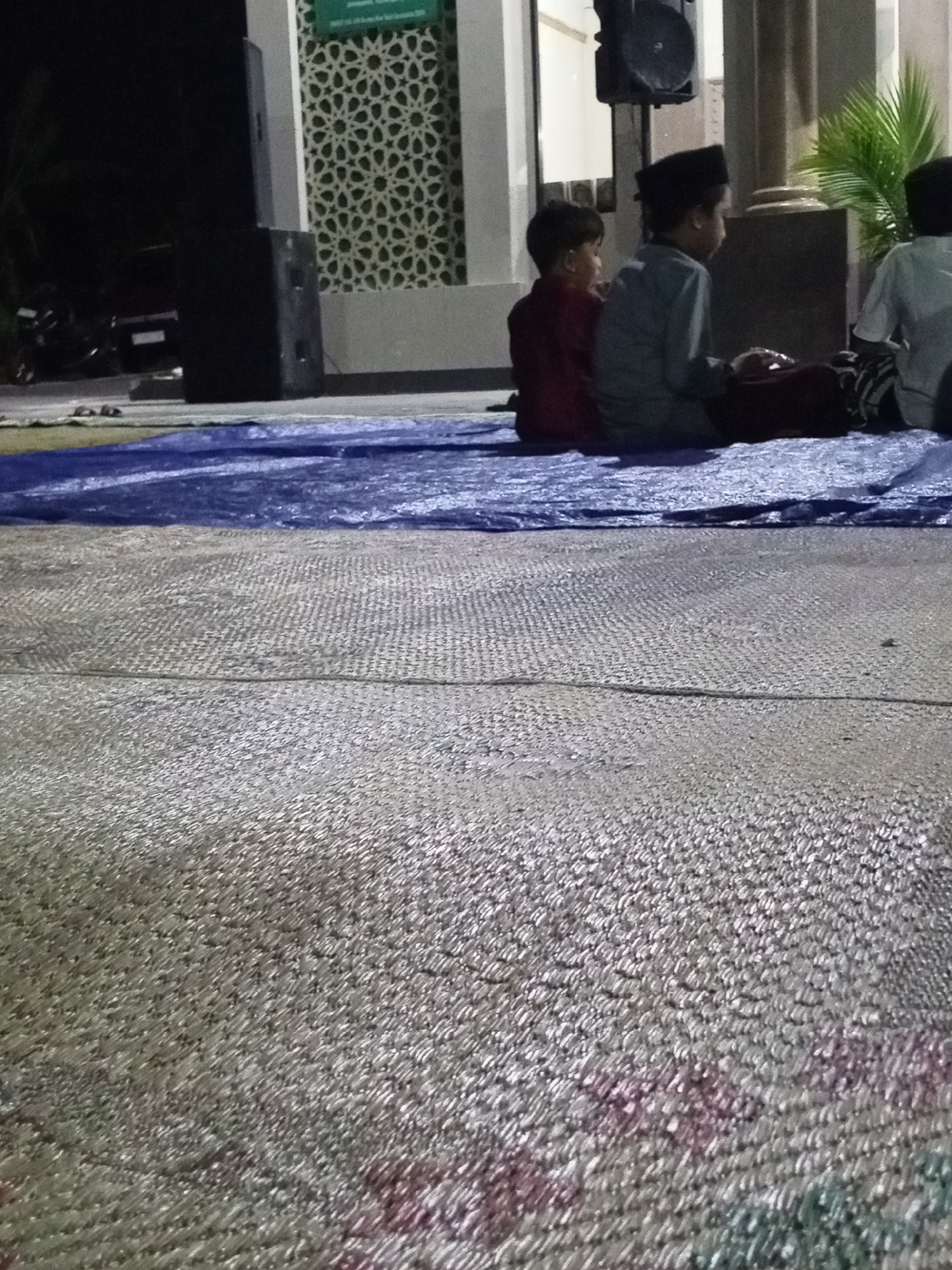 In Lemahbang, Indonesia, three young boys sit on a blue tarp spread on a textured surface, likely woven matting. The boy on the far left wears a dark red shirt. The middle boy, wearing a black peci and a light blue shirt, is in the process of praying or meditating. The boy on the far right, partially visible, wears a white shirt. Behind them, a large black speaker unit is mounted on a stand. Further back, a building with a distinctive patterned wall and a green sign is visible. The overall scene suggests an outdoor gathering, possibly religious or communal, taking place near a building that resembles a mosque or community center.