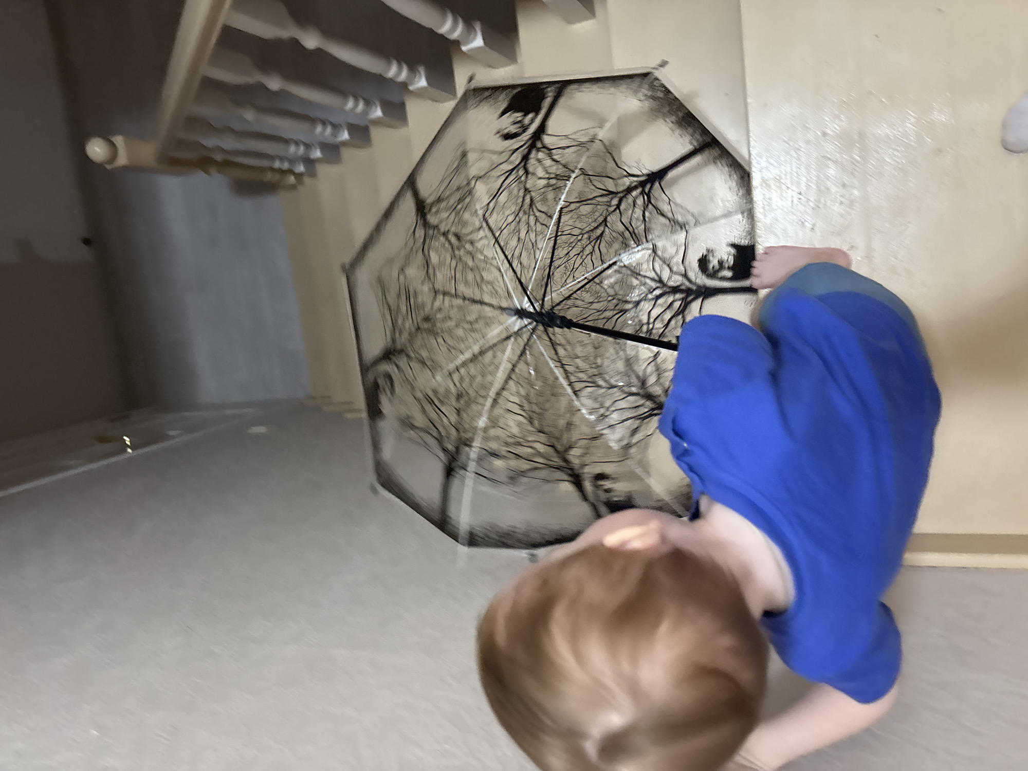 A child wearing blue pajamas stands on a light-colored floor next to an open umbrella with a tree branch pattern. A staircase is visible in the background. The location cannot be confirmed as being in Ферма, Russia.