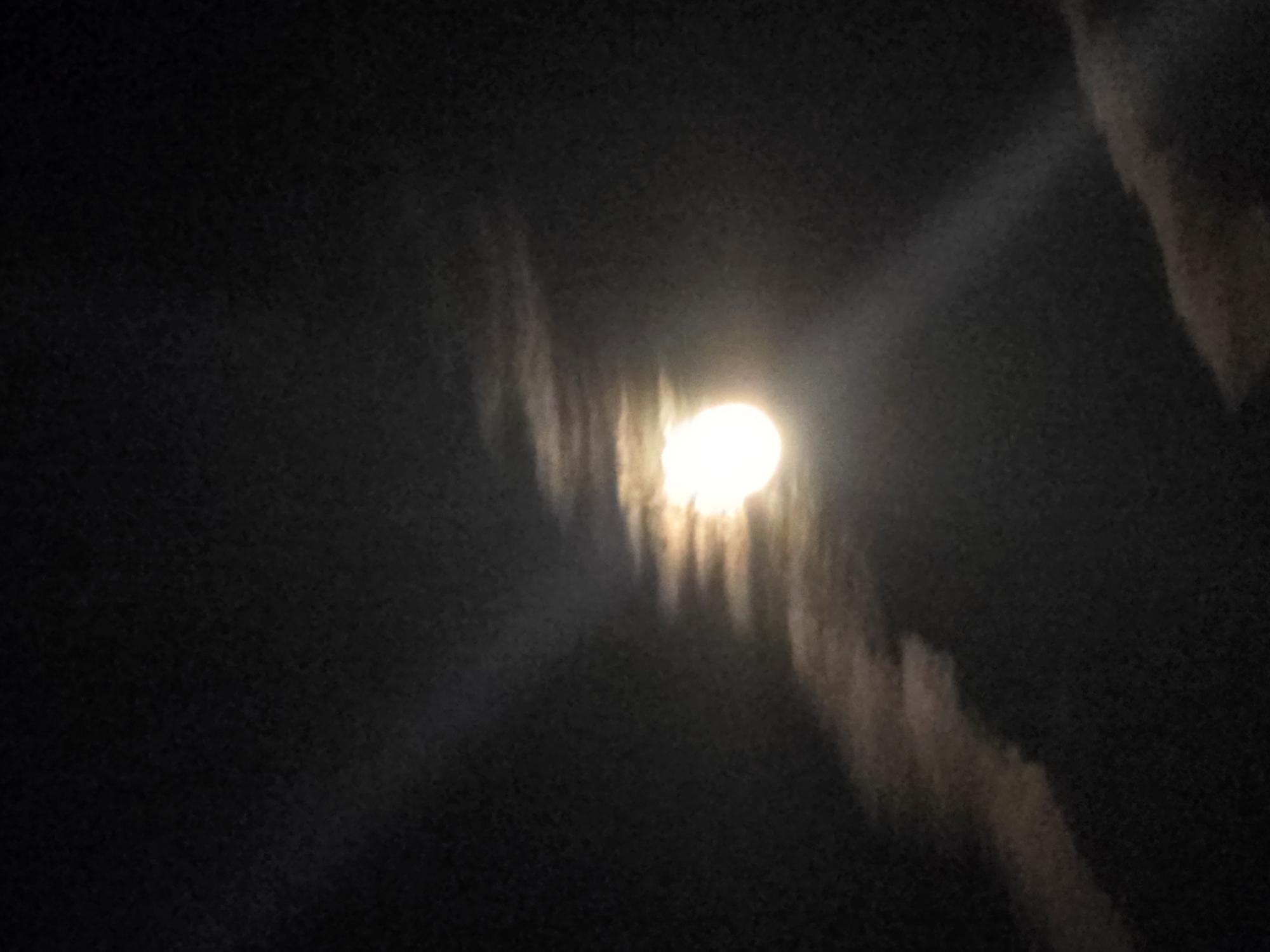 The image shows a full moon partially obscured by clouds at night. The moon is bright and appears to be the primary light source in the scene. Wispy clouds are visible, some catching the moonlight and appearing illuminated. The background is dark, indicating a nighttime sky. There are no people or man-made objects visible in the image. The image is a close-up shot focused on the moon and surrounding clouds.
