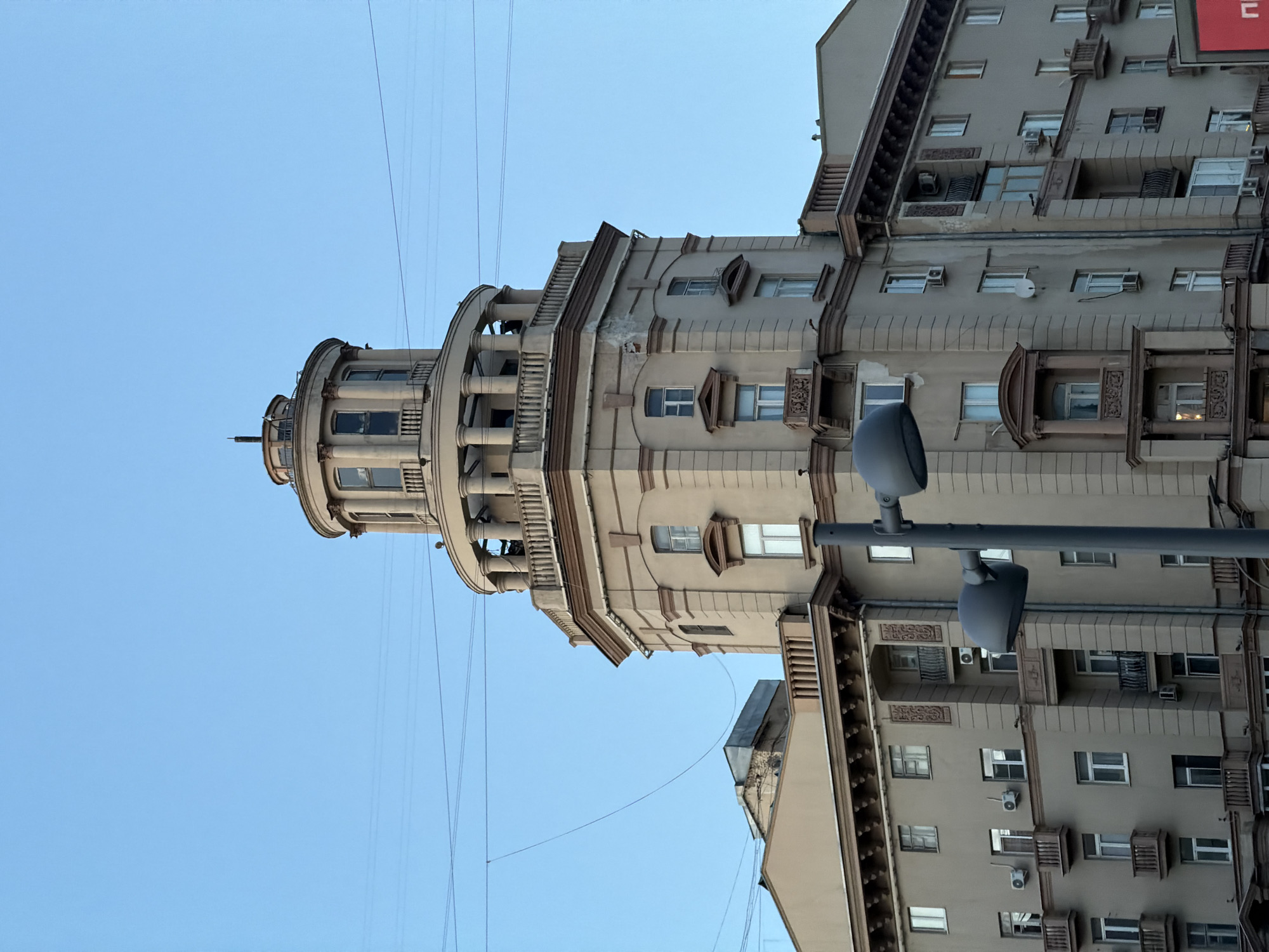 An image, captured in portrait orientation, depicts a grand, multi-story building against a clear blue sky in Moscow, Russia. The building, likely an apartment or mixed-use structure, is a light beige or cream color with darker brown decorative elements. Its architecture suggests a classical or Stalinist Empire style, featuring ornate cornices, decorative window surrounds, and intricate balcony railings.
A prominent feature is a rounded corner tower or turret on the left side, which rises above the main building. This turret has multiple levels, large windows, and columns, culminating in a small spire or finial at its peak. Numerous windows, many equipped with air conditioning units and some with satellite dishes, are visible across the building's facade.
In the foreground on the right, a grey street light pole with two horn-shaped speakers or lights extends into the frame. Several thin utility wires crisscross the expanse of the bright, cloudless blue sky.
The scene is set during the daytime, under bright, clear weather conditions. No people are visible. On the far right edge of the main image, a partial red sign with a white Cyrillic letter, possibly "Ц" or "П", is visible.