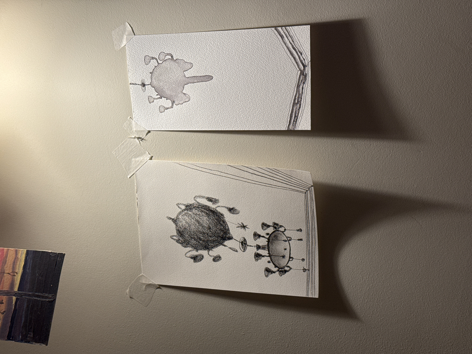 Two drawings are taped to a light gray wall. The upper drawing depicts a blob-like form with tendrils emanating from it, and parallel lines along the right edge. The lower drawing features two fantastical, creature-like forms connected by a thin line. One is rounded and dark, and the other more oval-shaped with spiky projections. To the left of the drawings, a portion of a painting with dark and colorful hues is visible. The lighting casts a shadow of the lower drawing onto the wall to its right. There is no information about the location in the city of Moscow, Russia in this image.