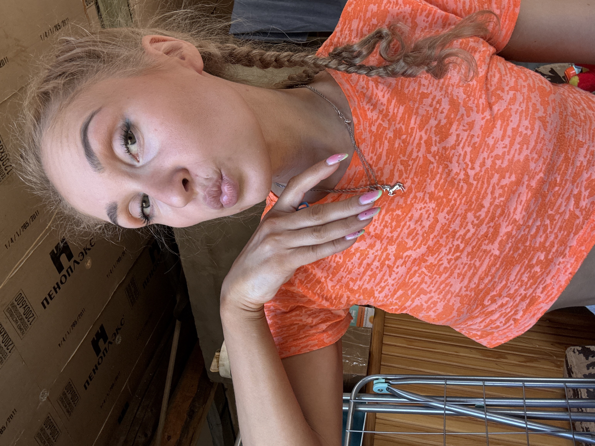 A young, fair-skinned woman with braided blonde hair and pouty lips, wearing a pink and orange patterned t-shirt and a silver horse pendant necklace, poses indoors. Her nails are painted with a pink gradient and green tips. The background includes stacked boxes with the brand "ПЕНОПЛЭКС" printed on them, a clothing rack, and wooden surfaces. The text on the boxes includes "1/4/1/285/2011" and "1200x600," indicating product dimensions. Location is indicated by the presence of Cyrillic text, which would place it within Russia. Specifically the Cyrillic logo of Пеноплэкс would place it in Nizhnyaya Chermoda, Russia.