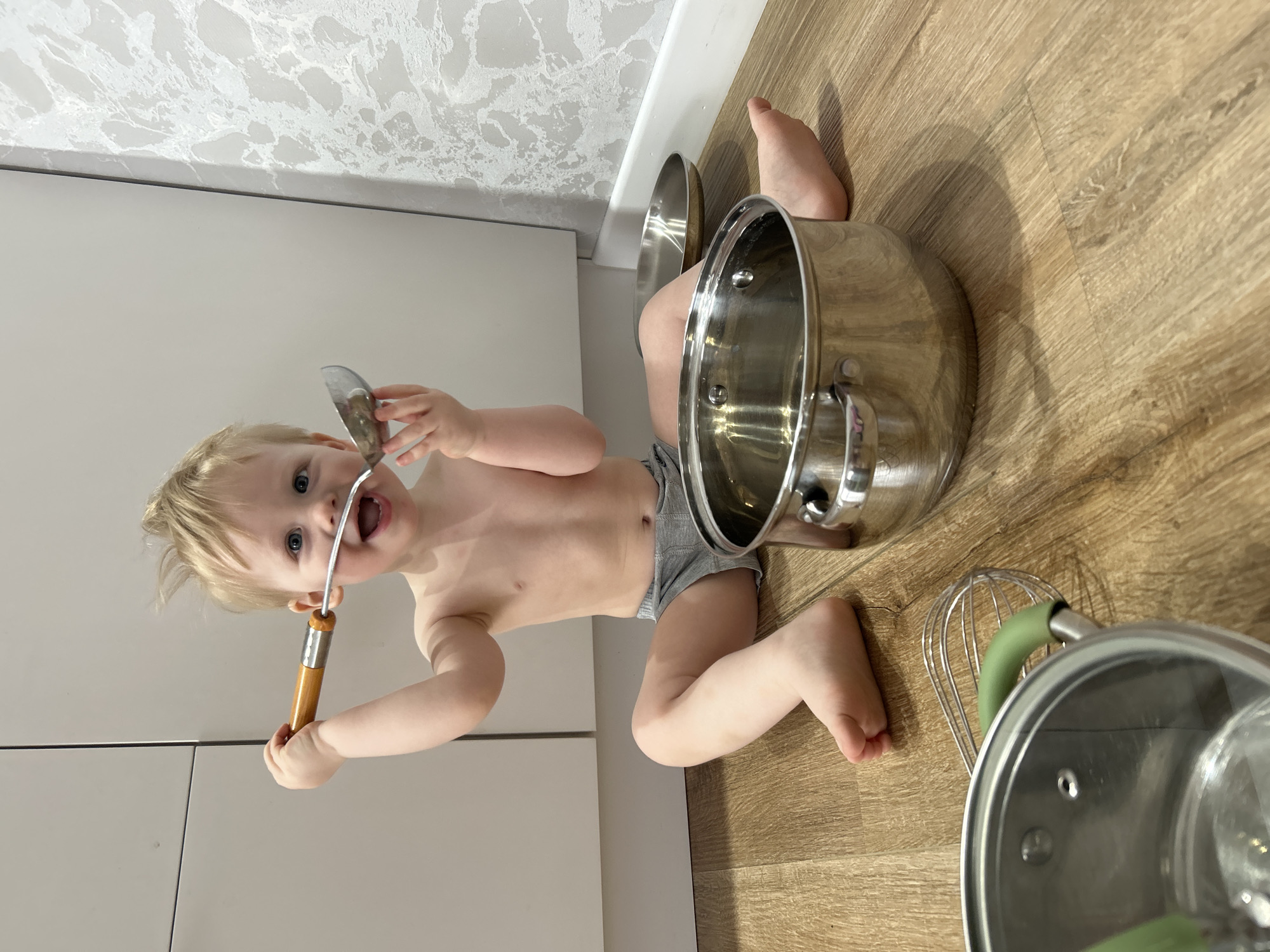 A young child is playing with kitchen utensils on a wooden floor in an interior setting. The child is fair-skinned, with light hair and is wearing gray underwear. They are holding a metal spatula in their mouth and another in their hand. Surrounding the child are several stainless steel pots and a whisk. The background consists of a textured wallpaper and light-colored cabinets. The location is in the city of Chaikovsky, Russia.