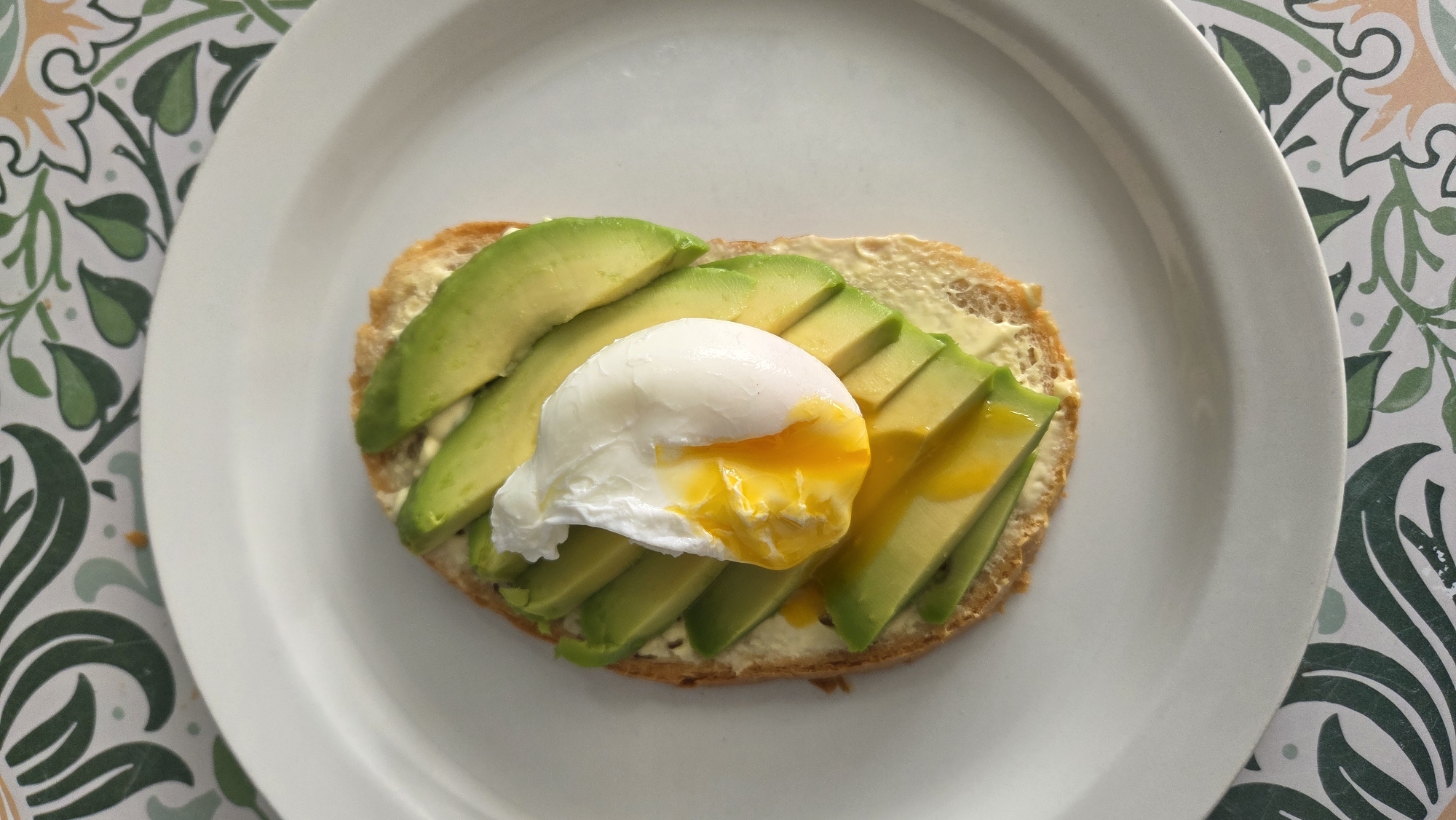 A single slice of toasted bread topped with sliced avocado and a poached egg sits on a white plate. The poached egg has a runny yolk that has spilled onto the avocado. The plate is on a patterned tablecloth with green and white leaf designs. The image is taken from a top-down perspective. There are no people or animals visible in the image, and there is no indication of the location being in Tynset, Norway.