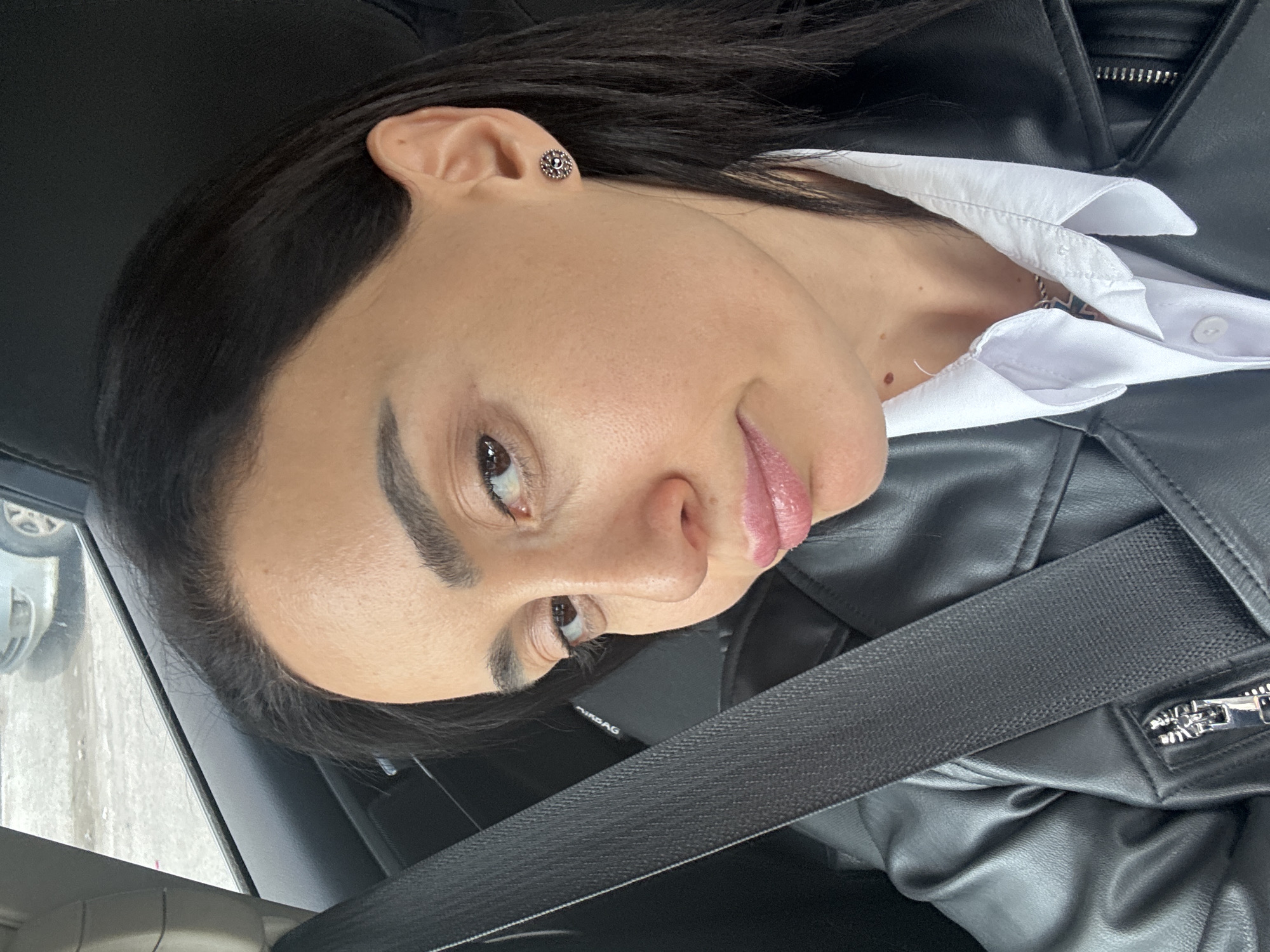 A woman is seated inside a car, wearing a black leather jacket and a white collared shirt. A seatbelt is fastened across her body. She has dark hair and is wearing an earring in her left ear. The image appears to be taken from a slightly high angle, looking down at her. The location appears to be in Tbilisi, Georgia. The background shows a blurry view through the car window, including a light-colored vehicle and what seems to be an outdoor environment, likely urban.