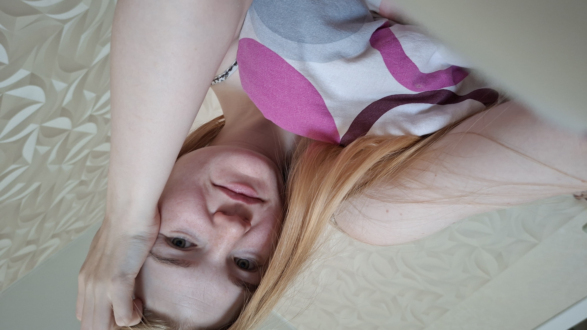 The image shows a close-up, overhead shot of a young woman with blonde hair and fair skin with freckles. She is lying down, propped up on her arm, with her head resting on her hand. Her eyes are open and looking towards the camera. She is wearing a top with a pink and purple abstract pattern. The background appears to be a wall with a textured, light-colored wallpaper. There are no discernible city landmarks or specific details that would indicate a location in Chaikovsky, Russia, or any other city. The scene is intimate and personal, suggesting a casual, indoor setting.