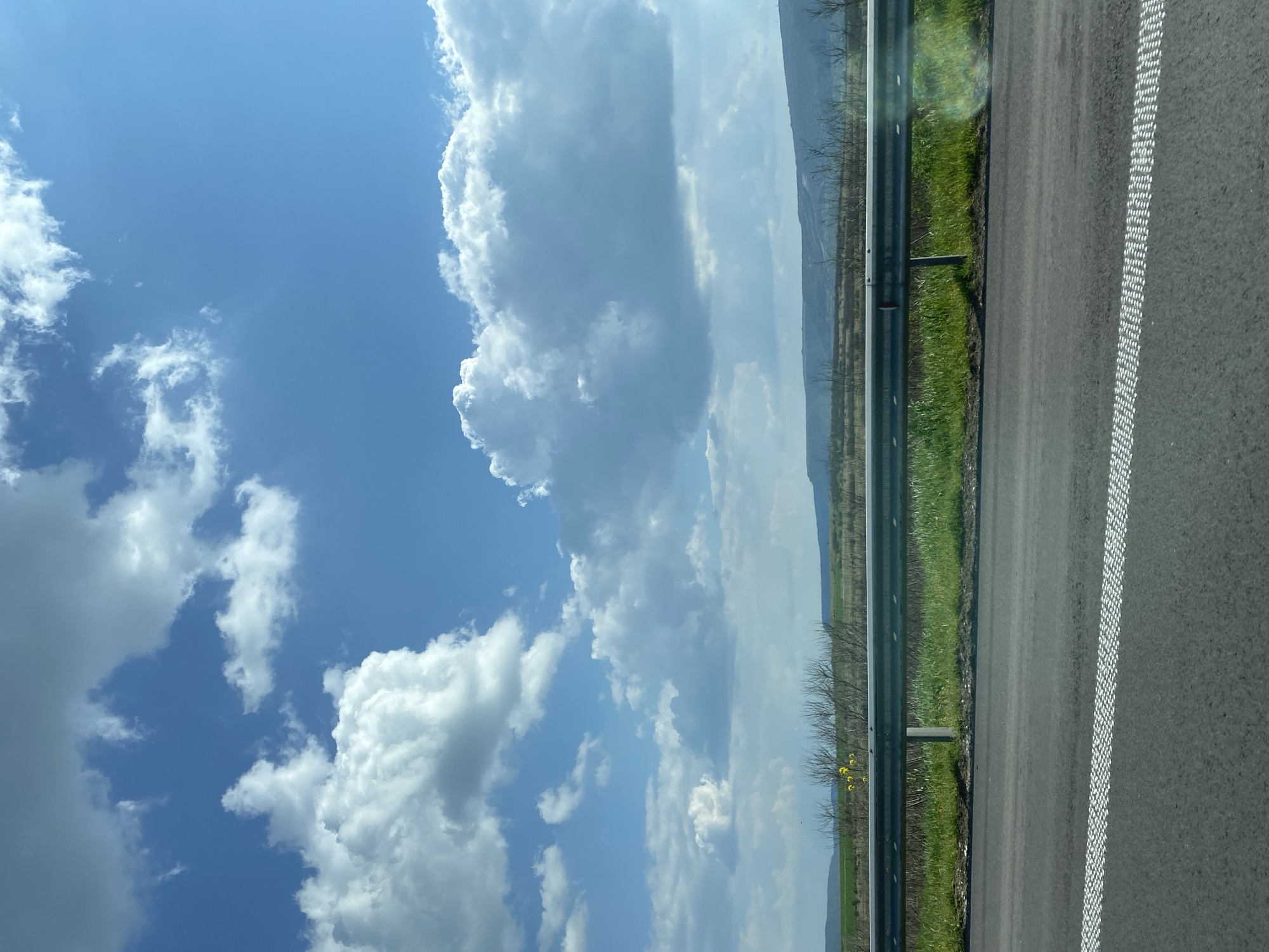 The image shows a view from a vehicle traveling on a road, likely a highway, in or near Zlatina, Bulgaria. The right side of the frame is dominated by the road surface, with white dashed lines marking the edge of the lane. Beyond the road is a grassy shoulder, a guardrail, and a landscape with trees and what appears to be a distant ridge or mountain range. The left side of the frame displays a bright blue sky with puffy white clouds, which occupy a significant portion of the image. The overall composition indicates a sunny day with clear visibility. There are no people visible in the image.