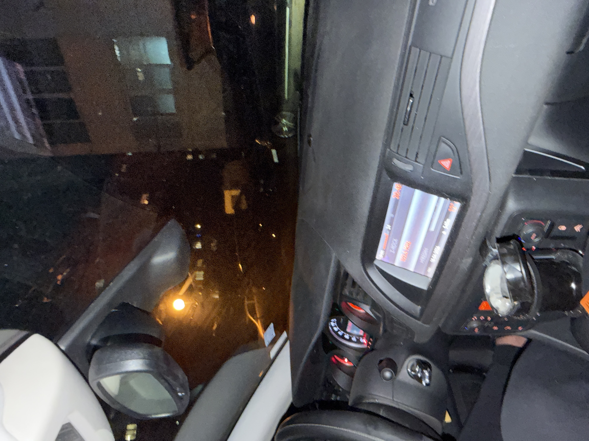 The image depicts the interior of a car, likely taken at night, with a view through the driver's side window. The car's dashboard is visible, including the infotainment screen, climate control vents, hazard lights button, and instrument cluster. A side view mirror is also in view. Through the window, a cityscape is visible, suggesting an urban environment. The location is Sofia, Bulgaria, evidenced by the reflection of buildings and streetlights.