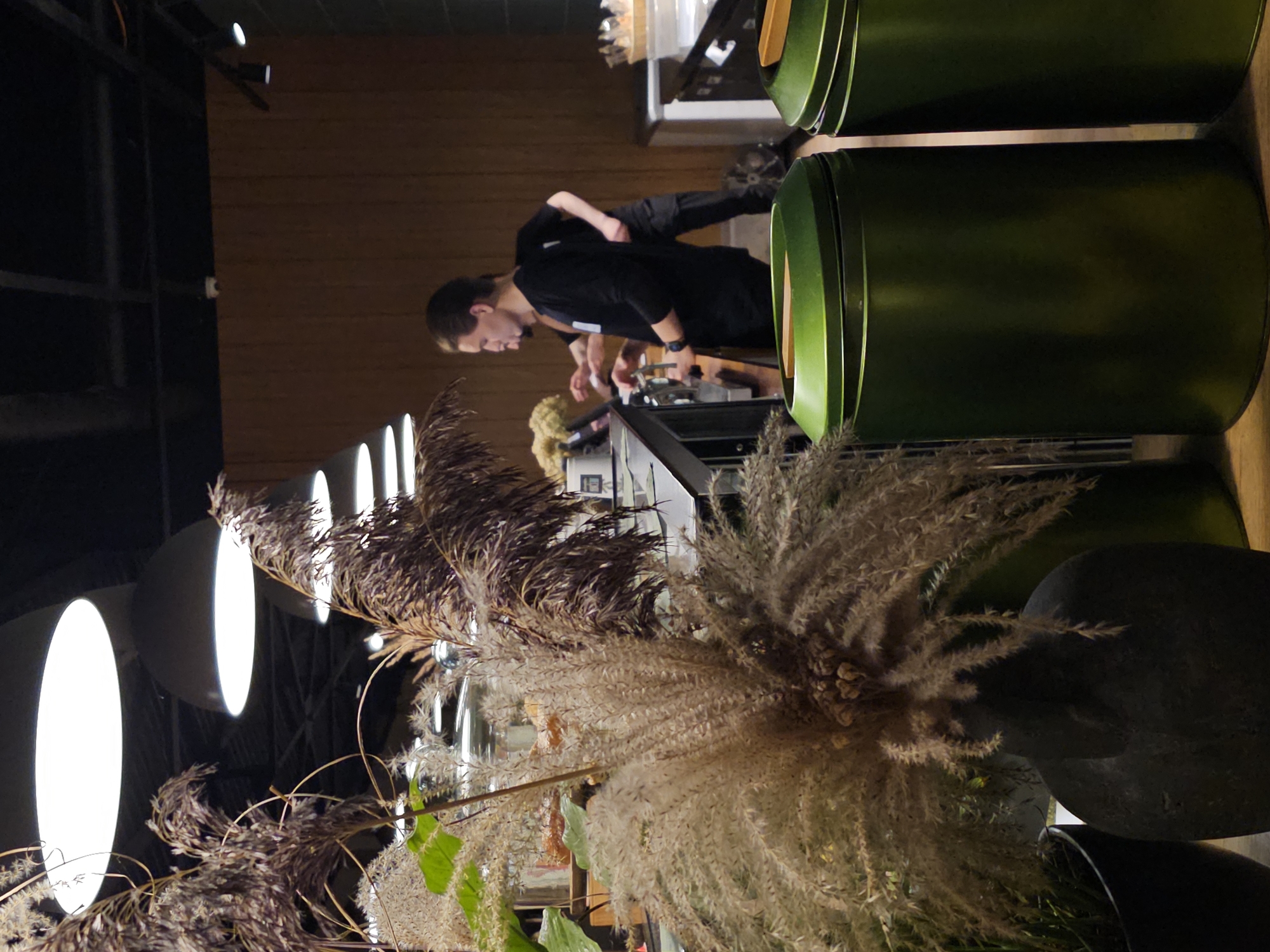 In Nemchinovo, Russia, this image captures an indoor scene, possibly a cafe or restaurant. The foreground is dominated by a large, dark, textured vase holding a substantial arrangement of dried grasses and reeds, with a hint of green foliage. To the left of the vase, a portion of a black planter is visible. In the middle ground, a counter area is present, with several people interacting behind it. One individual, wearing a black long-sleeved shirt and dark pants, is actively engaged with a coffee machine. Another person, also in dark clothing, stands behind them. The background features a warm-toned wooden wall and large, circular light fixtures suspended from the ceiling. In the lower right of the frame, two large, dark green cylindrical containers with wooden lids are prominent. The overall impression is of a contemporary establishment with naturalistic decorative elements.