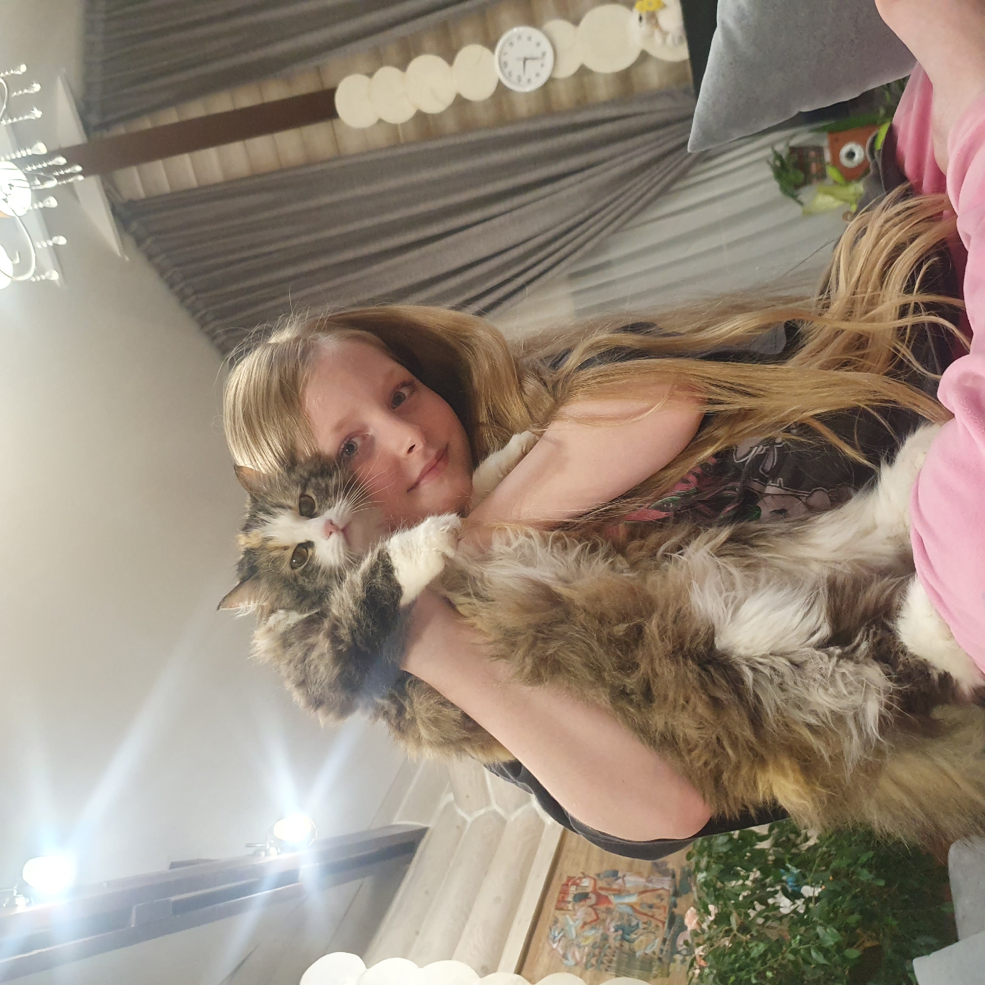 A woman with long blonde hair is holding a fluffy calico cat. The cat is resting in the woman's arms. The setting appears to be indoors, potentially a living room, and the background includes decorative elements like wall hangings and a clock. The image orientation is rotated. There is no indication of the location being Chaikovsky, Russia.