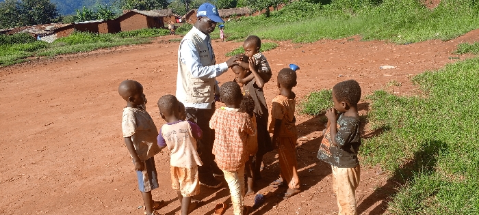 An outdoor scene in Ngalula, Democratic Republic of the Congo, shows one adult male and six young children on a reddish-brown dirt road under bright daylight. The adult male, wearing a light blue baseball cap, a light blue long-sleeved shirt, and a multi-pocketed beige vest with "WFP" visible on the left chest, stands centrally, facing the children. His right hand is placed on the head of a child standing in front of him, who is wearing a dark patterned top. A toddler, dressed in a dark top, is held by the adult, facing away from the camera. Four other children, clad in various casual tops and pants, are positioned around the adult, observing the interaction. A child to the left wears a light-colored top and striped shorts, while a child to the far right wears a dark patterned top and light brown pants, with hands near their mouth, standing near a blue plastic container on the ground. The unpaved road displays faint tracks. Green grass and low bushes flank the road's edges. In the background, several single-story, mud-brick or concrete block structures with tin roofs are visible amidst sparse trees and vegetation, against a backdrop of a tree-covered hillside.