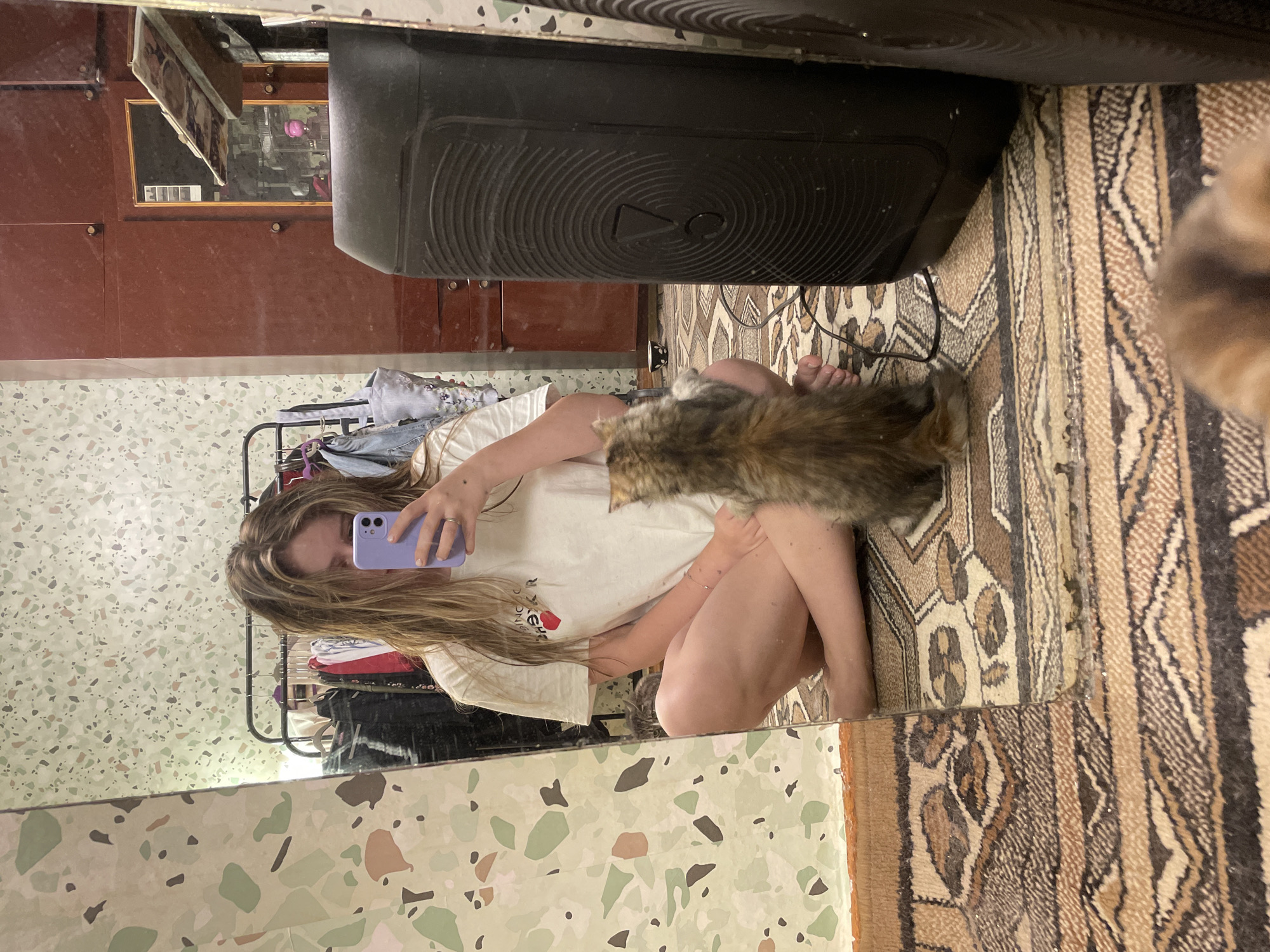 A young woman is sitting on a patterned rug, legs crossed, facing a mirror. She is holding a purple smartphone in her right hand, taking a selfie. Her left hand rests on her lap, and she wears a white t-shirt with a graphic design on the front. A small, fluffy cat is sitting on the rug in front of her, looking up at her. The background is a room with a multi-colored speckled wallpaper and a clothing rack with clothes hanging on it. To the right of the woman, a large black speaker is visible. Another cat's face is partially visible in the foreground, out of focus.