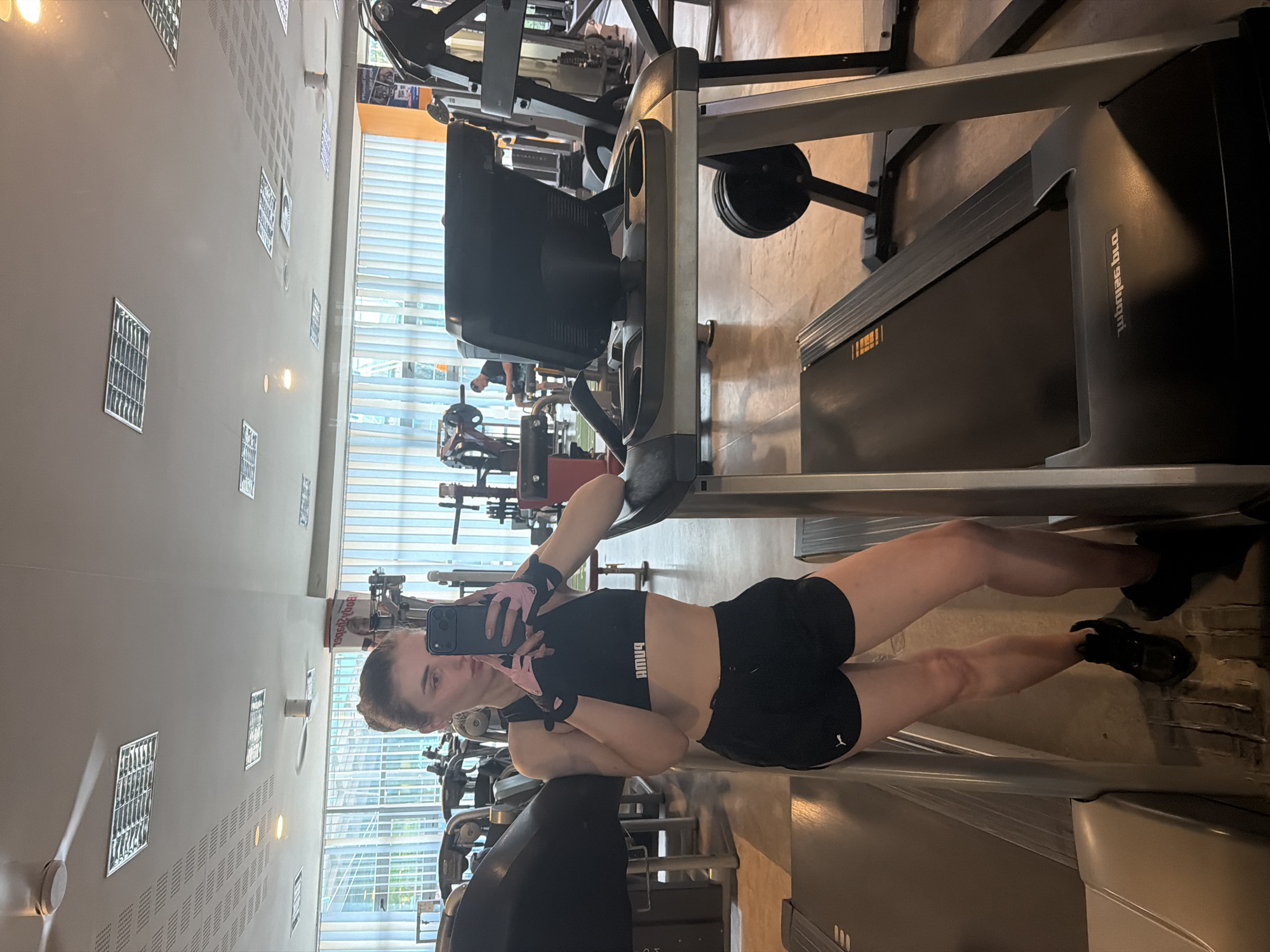 A young woman with blonde hair tied up in a bun is taking a mirror selfie in a gym. She is wearing a black sports bra, black shorts, and black athletic shoes with pink wrist wraps. She is holding a black smartphone in her right hand and leaning on a treadmill with her left arm. The treadmill is a dark gray color with a black belt and a display console. In the background, other gym equipment, including treadmills and weight machines, are visible, as well as large windows with vertical blinds. A man is seated on a weight machine in the background. The gym appears to be well-lit with fluorescent lighting.