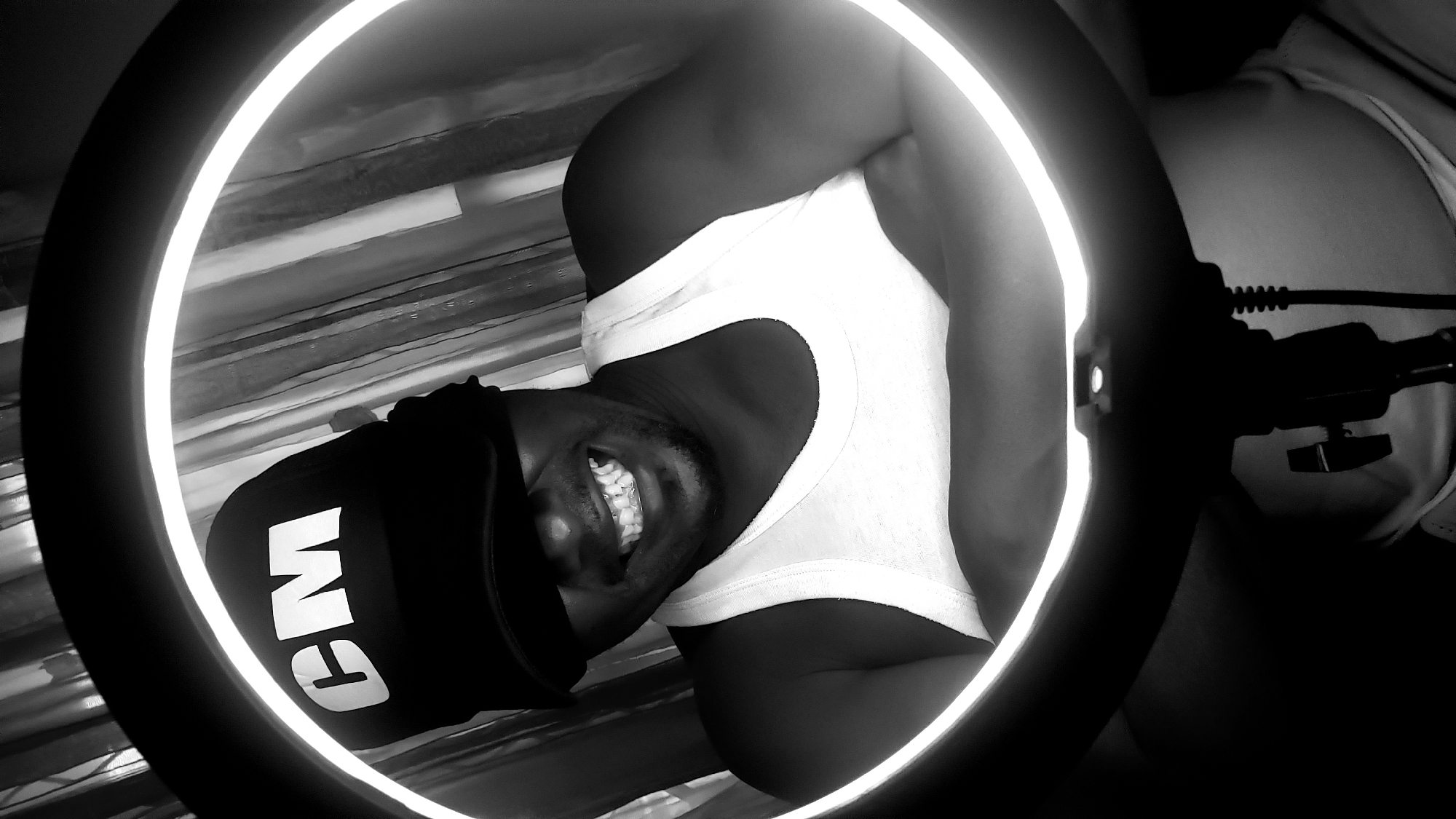 A black and white portrait shows a dark-skinned person smiling, with teeth clearly visible. The person is wearing a white tank top and a black baseball cap with "CM" in large white letters. The person is illuminated by a ring light, with the light's mount and a cord visible on the right side of the image. A wall with horizontal patterns appears in the background. The image does not contain enough information to confirm the location in Sabon Tasha, Nigeria.
