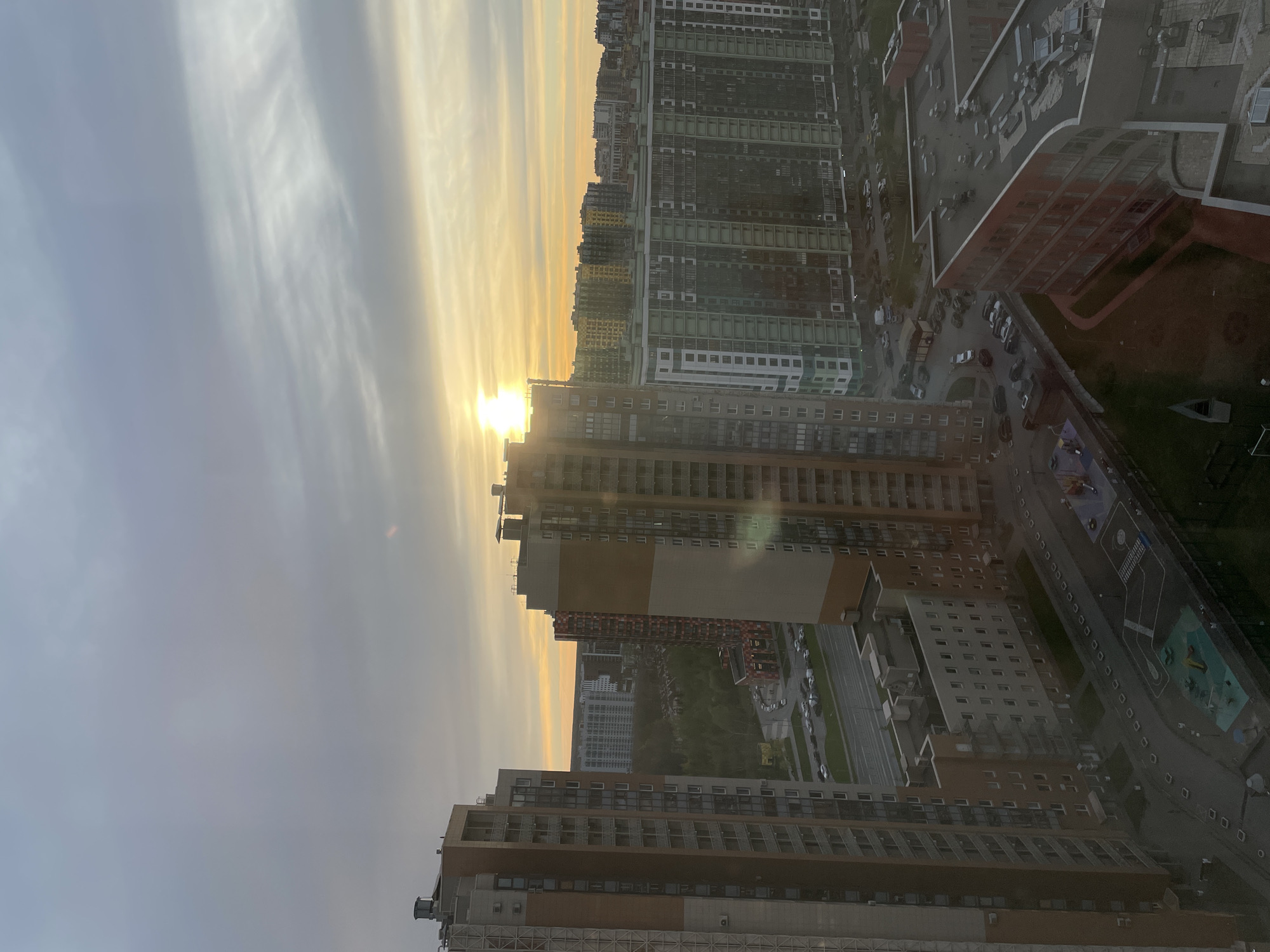 The image shows an urban scene in Saint Petersburg, Russia, at what appears to be sunset or sunrise. The sun is low on the horizon, obscured by clouds, casting a golden glow over the city. The view is from a high vantage point, likely an upper floor of a tall building, looking down at a dense collection of apartment buildings of varying heights and colors, including brown, beige, green, and red. A park or green space is visible between some of the buildings. Cars are parked on streets surrounding the buildings. A playground area is discernable amongst the buildings on the right side of the image. The sky is a mix of grey and golden colors, suggesting a cloudy day with sunlight breaking through.