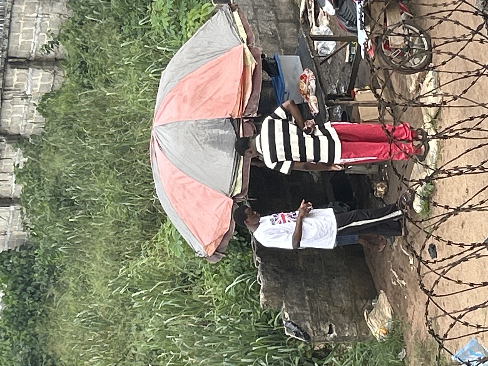 In Ihiagwa, Nigeria, the scene reveals two individuals standing beneath a weathered, two-toned umbrella, providing shelter from the sun. One individual wears a white t-shirt with an emblem and black pants, while the other wears a black and white striped shirt and red pants. The area is overgrown with dense green vegetation. A makeshift shelter is constructed nearby, cluttered with various items, including a bicycle wheel. Barbed wire is coiled in the foreground, adding a sense of enclosure to the scene.