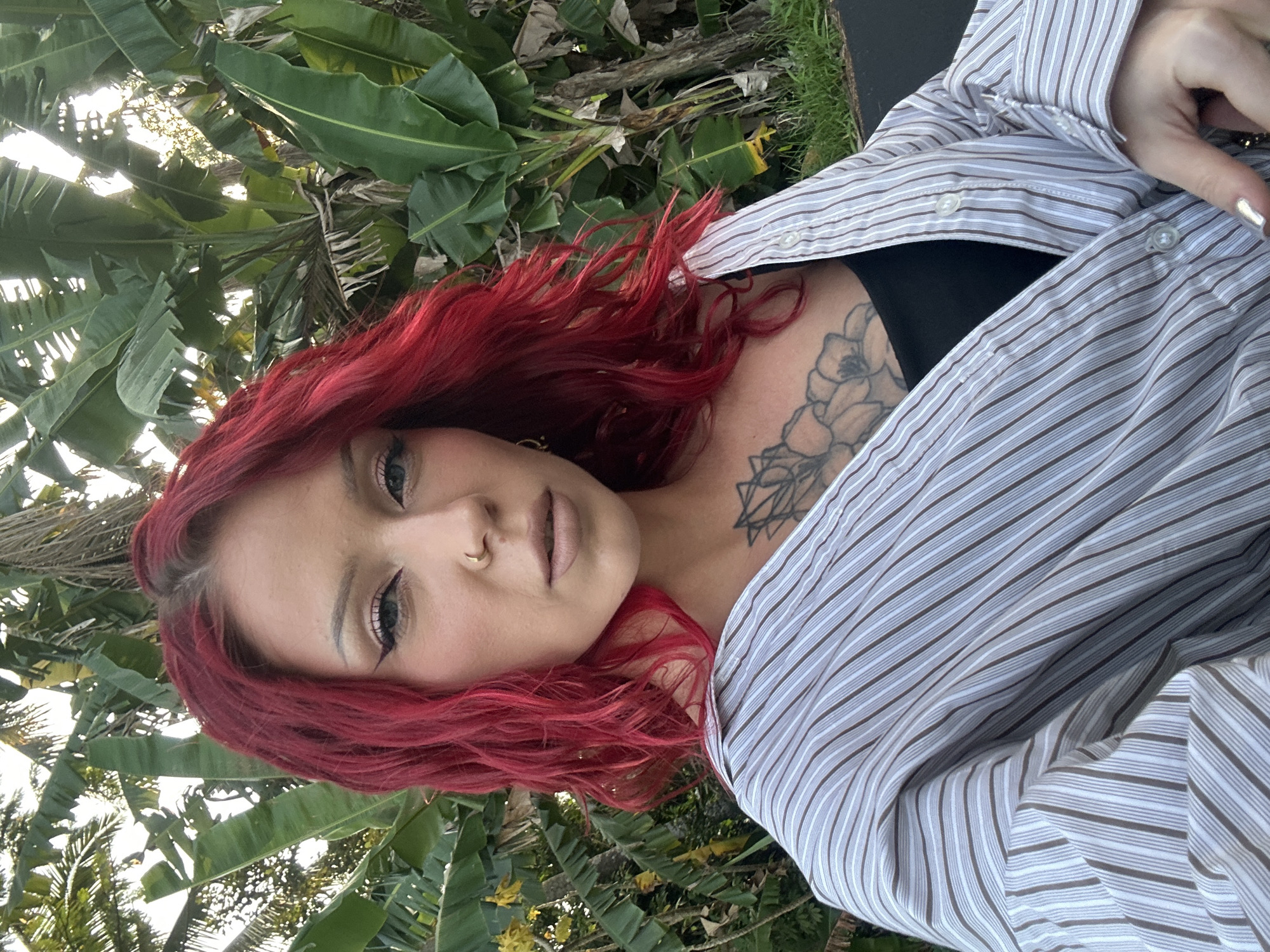The image features a person with vibrant red, wavy hair, looking directly at the camera with a neutral expression. They have fair skin, dark winged eyeliner, a nude lip, and a gold nose ring. A detailed black-ink floral tattoo is visible on their upper chest and collarbone area.
The individual is wearing a light-colored, striped button-up shirt with thin brown and grey vertical stripes, worn open over a black top. Their left hand, adorned with light-colored nail polish, is seen in the upper right portion of the frame.
The background is a dense, lush tropical environment dominated by large green leaves, characteristic of banana trees and possibly palm fronds. Bright sunlight filters through the foliage, creating illuminated spots and indicating a sunny day. Patches of green grass are also visible. The setting suggests an outdoor, natural landscape, consistent with a tropical location like Ubud District, Indonesia. No visible text is present in the image.