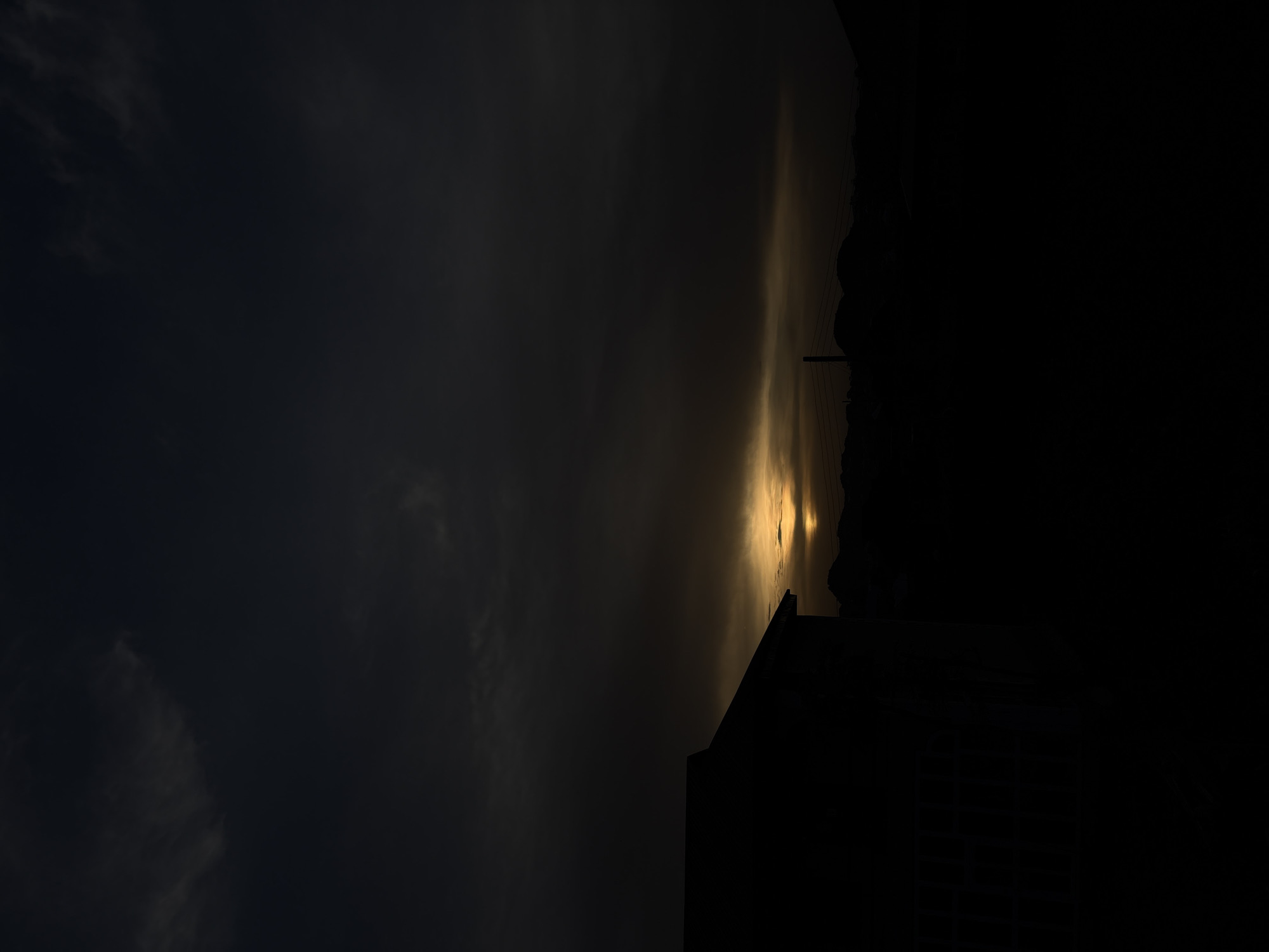 This image, taken in Bukuru, Nigeria, captures a dramatic sky scene at either sunrise or sunset. The sky dominates the frame, showcasing a transition from dark, heavy clouds in the upper and left portions to a brilliant golden-orange glow along the horizon on the right. This intense light, likely the sun partially obscured by clouds or haze, illuminates the lower sky and casts strong silhouettes across the landscape.
In the foreground, deep shadows render the details of the terrain and structures largely indistinct. To the left, the dark silhouette of a building's roofline and upper section is visible against the darker sky. Towards the center and right, the prominent profiles of mountains or hills rise against the bright horizon. A utility pole with power lines is also silhouetted against the golden light, indicating human infrastructure within this natural setting.
The scene is devoid of people or discernible activity. The heavy cloud cover contributes to the dramatic contrast between the dark skies and the vivid low-angle sunlight. No visible text is present. The overall mood is one of serene natural grandeur, highlighted by the interplay of light and shadow at the edge of day.