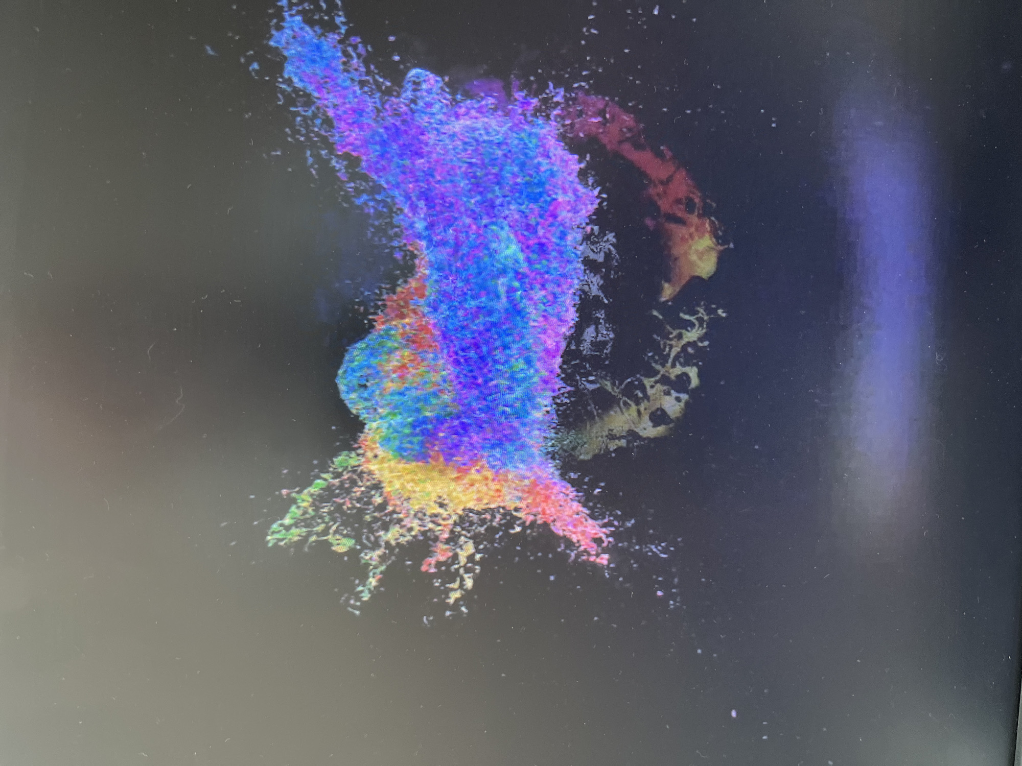 The image displays a multi-colored, abstract, cloud-like formation against a dark background. The colors range from blue and purple at the top, transitioning to yellow, orange, and pink towards the bottom. The formation has a nebulous, dispersed appearance, with small particles and streaks extending outwards. The context is unknown, and there are no identifiable people, subjects, objects, or actions to describe further. The location is unknown.